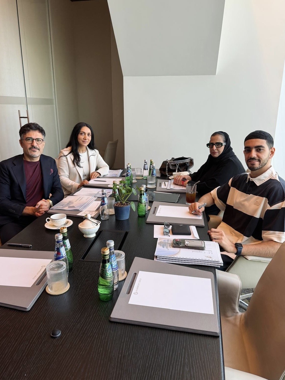 Fünf Menschen sitzen bei einem Meeting um einen Tisch in einem modernen Konferenzraum. Es gibt Getränke, Notizblätter, Laptops, und eine Pflanze auf dem Tisch.