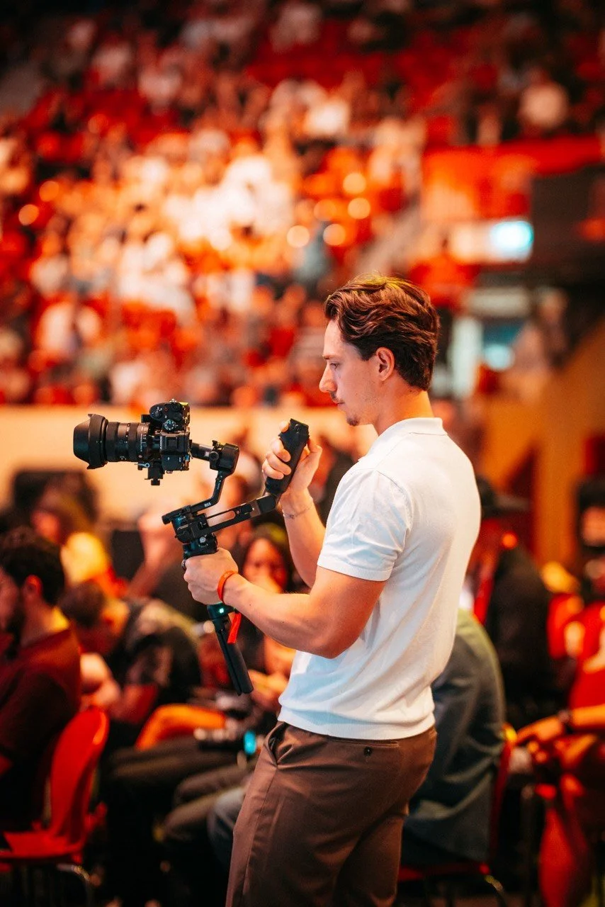 Young man in a white shirt holding a camera stabilizer and a microphone, standing in a crowded indoor event with a blurred audience in the background.
