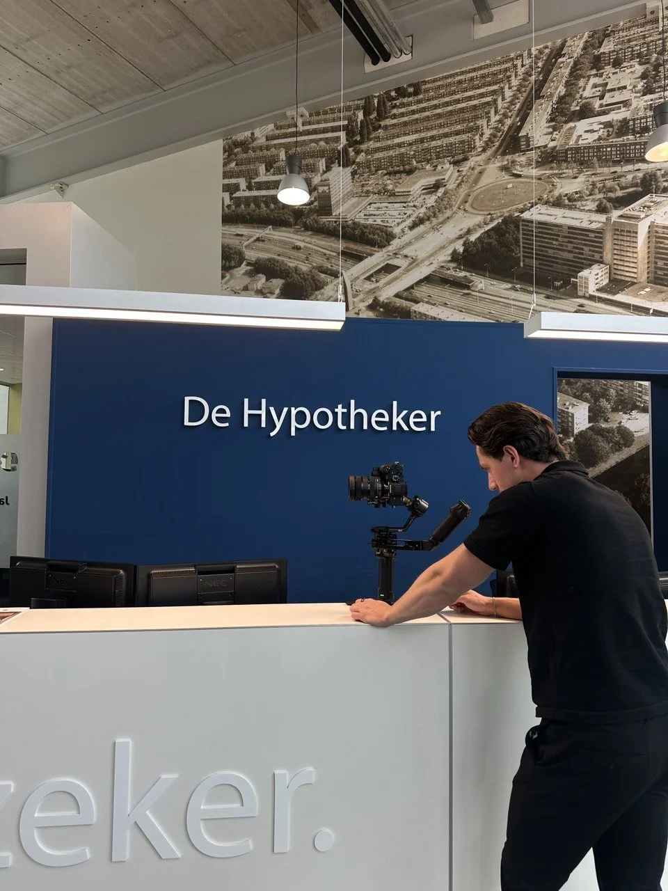Man setting up camera on a white counter at De Hypotheker, a mortgage company, with a blue wall and city aerial photo in the background.