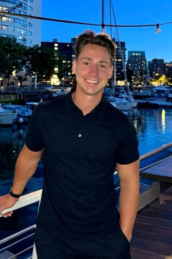 A young man with brown hair and a bright smile, wearing a black polo shirt and black pants, standing on a dock by a marina with boats, in an urban setting during dusk.