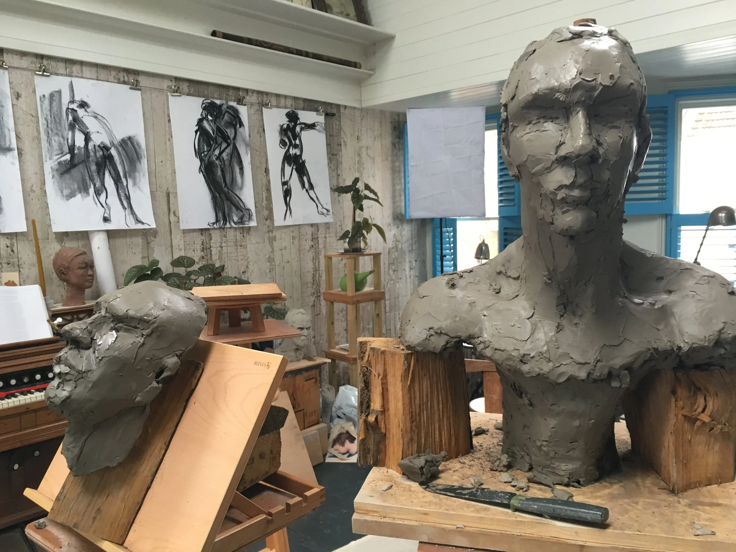 An art studio with various sketches and paintings on the wall, sculptures of human heads and figures made from clay, wood blocks, and art supplies scattered around.