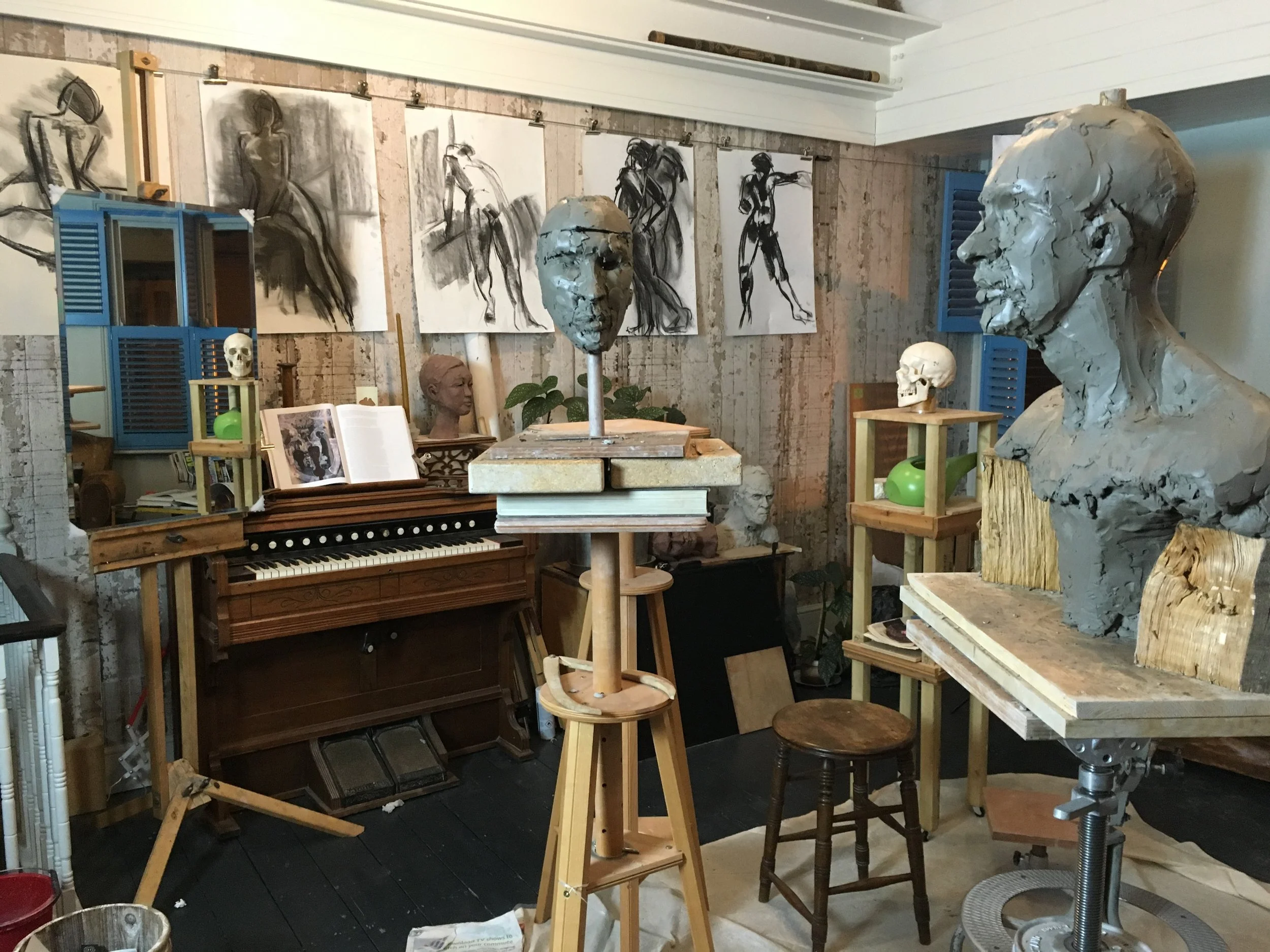 An art studio with sketches of female figures on the wall, bust sculptures, wooden stools, a piano, and various art supplies and sculptures.