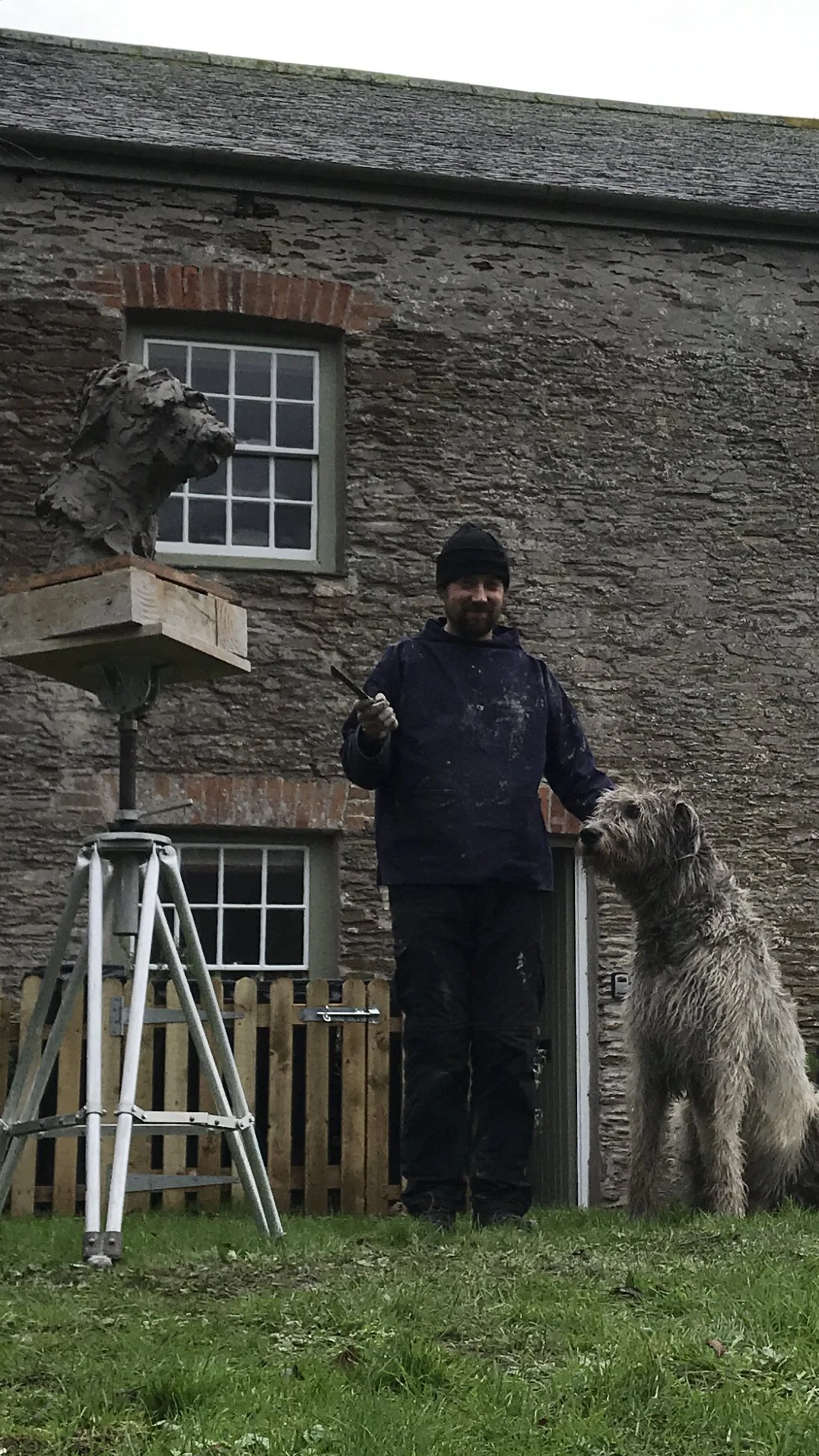 A man with a beard and black beanie stands outdoors next to a large, shaggy dog in front of a stone house. The man holds a paintbrush, and there is a sculpture of a dog's head on a platform nearby.