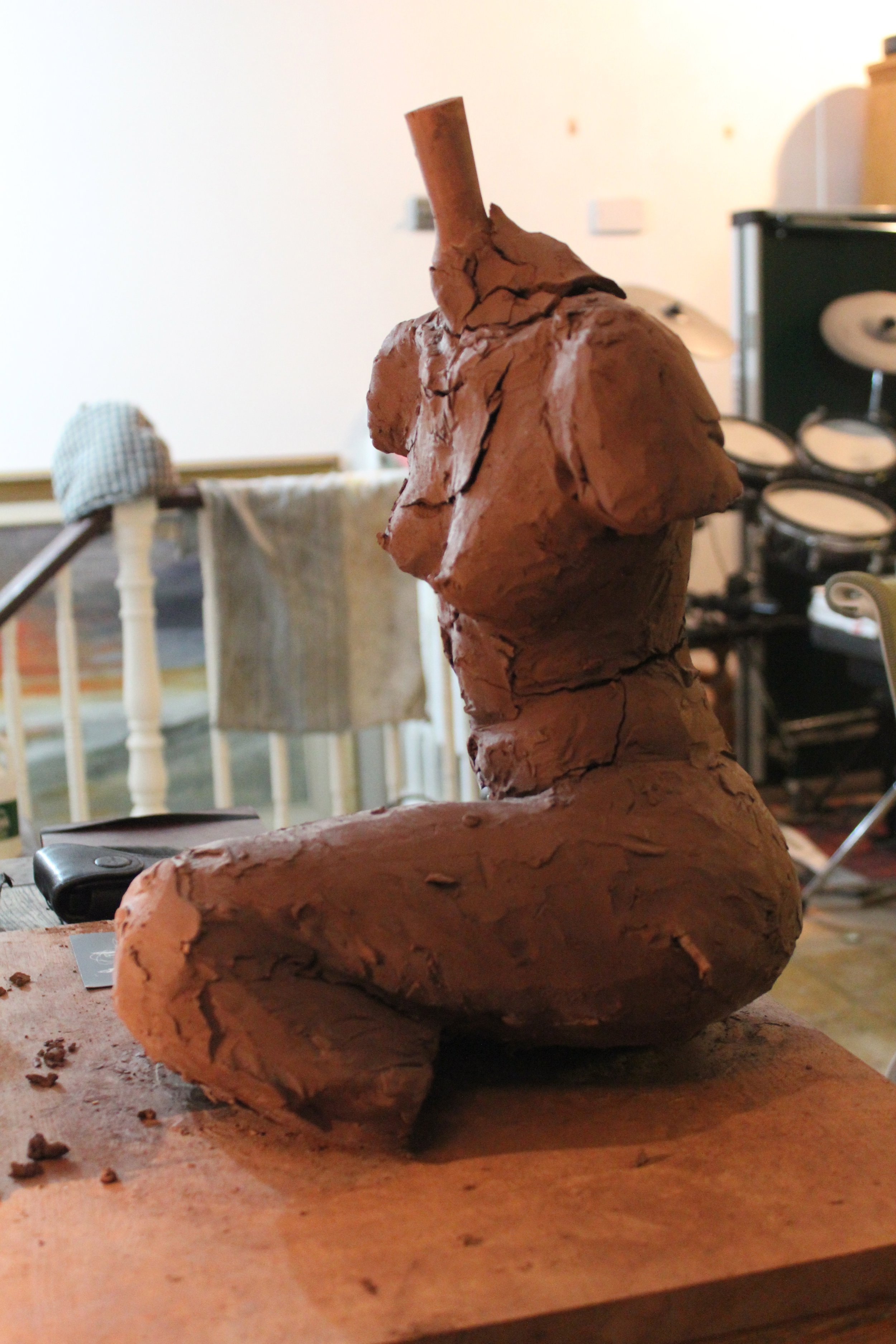 Close-up of a clay sculpture of a humanoid figure with prominent facial features, standing on a wooden surface in an indoor studio setting.