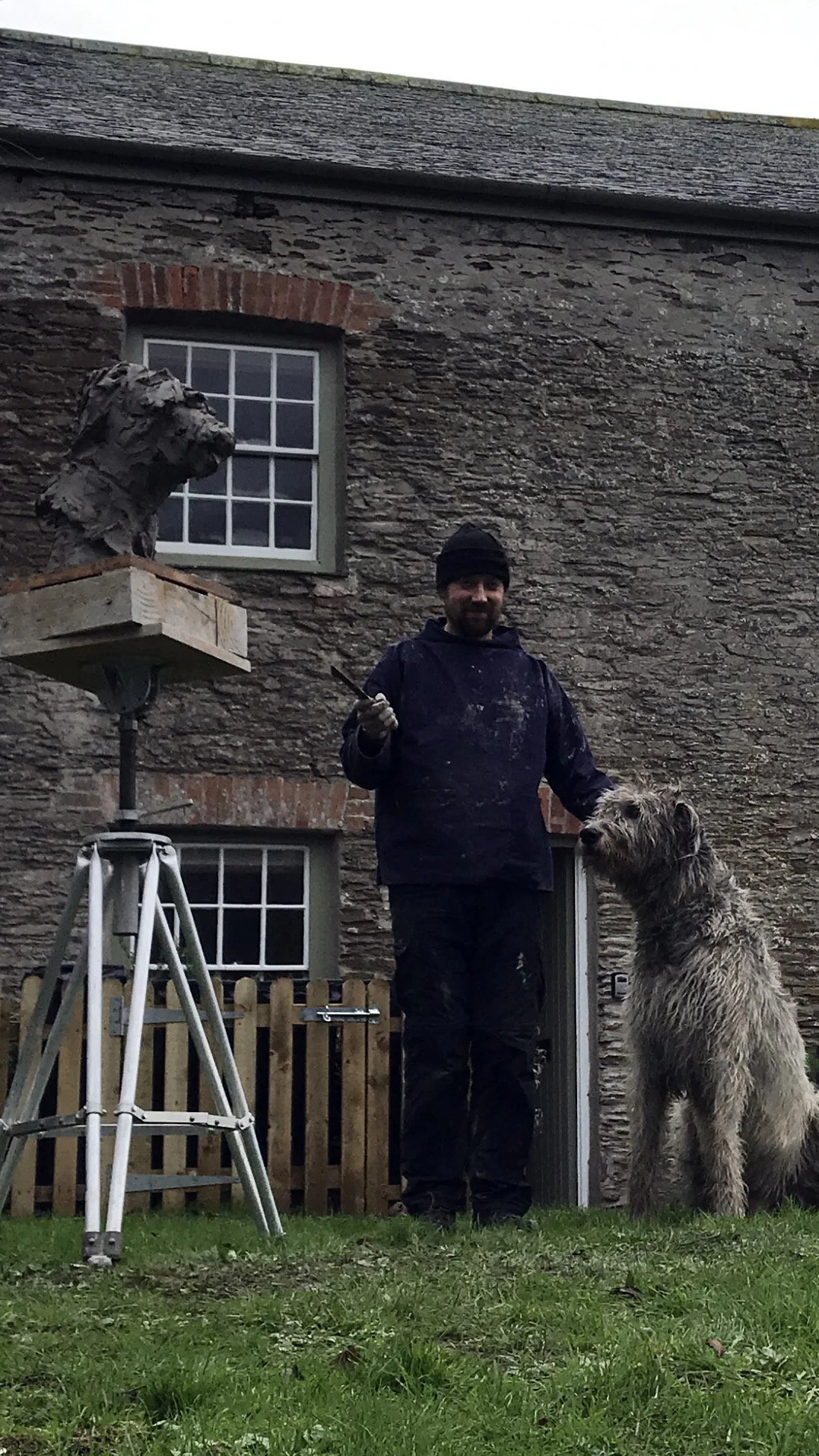 A man in black clothing and a beanie hat standing outside with a large shaggy dog at his side, holding a paintbrush. There is a building with stone walls and windows in the background, and a sculpture of a dog’s head on a pedestal next to a ladder.