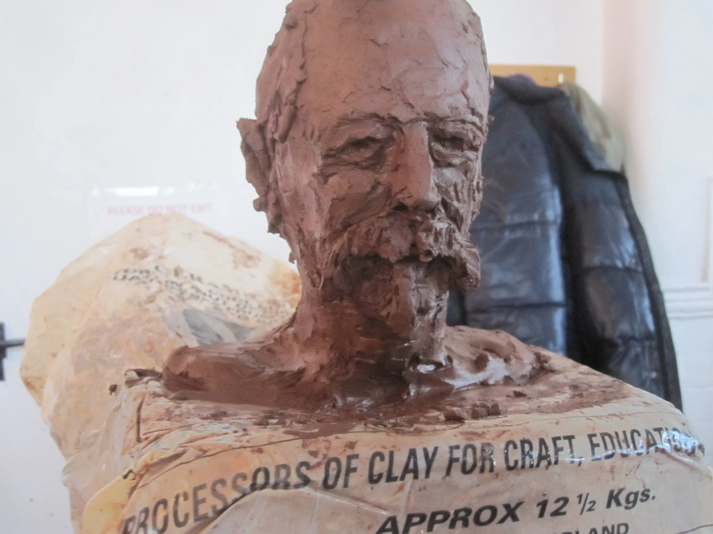 A sculpture of a bearded man's head made of clay, with a textured surface, mounted on a base with partially visible text about clay for craft and approximate weight.