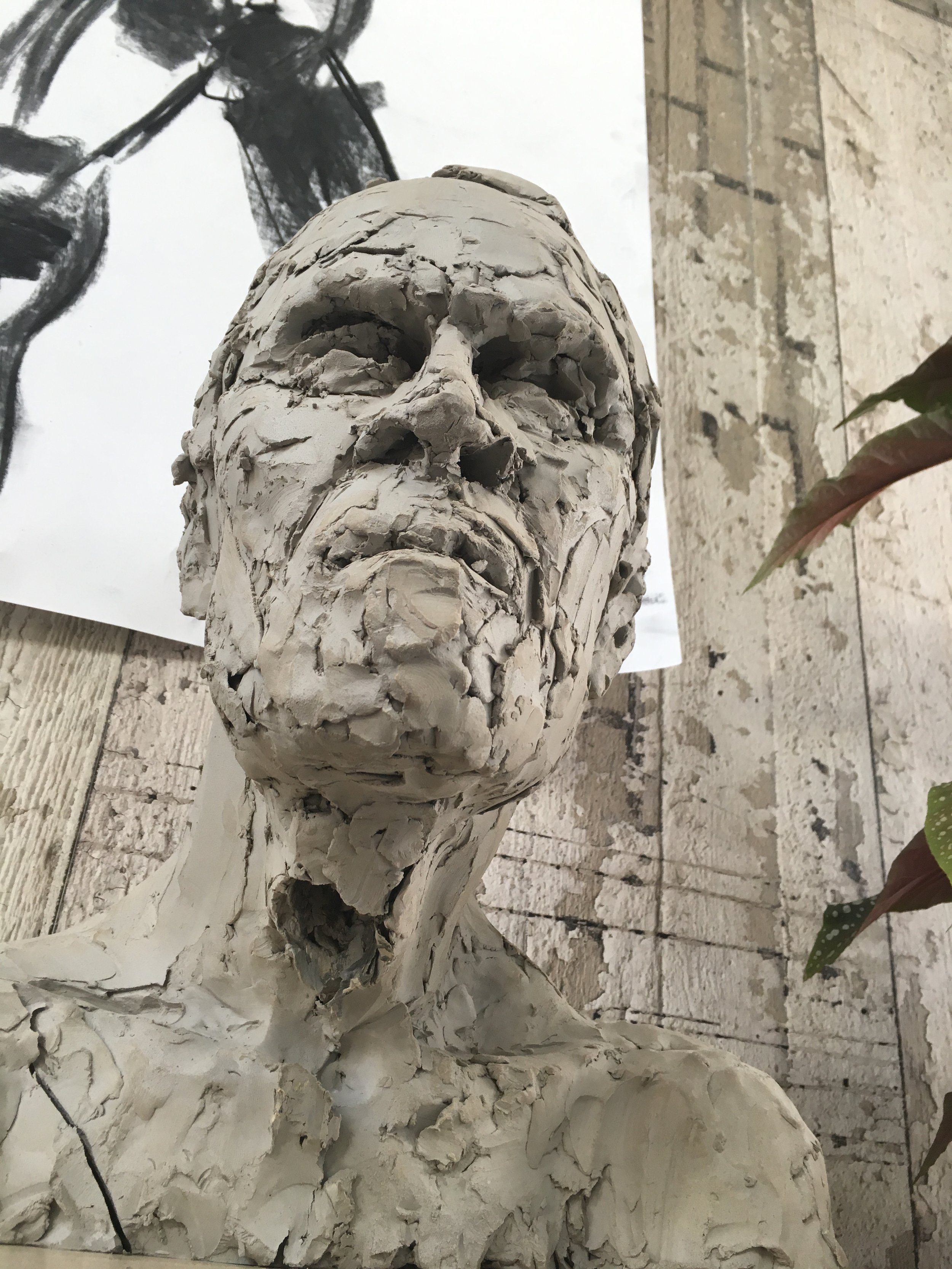 A white plaster bust sculpture of a person's head and shoulders with a rough, textured surface. The sculpture is placed on a flat surface, and behind it, there is a black and white abstract drawing on paper and a brick or stone wall background.