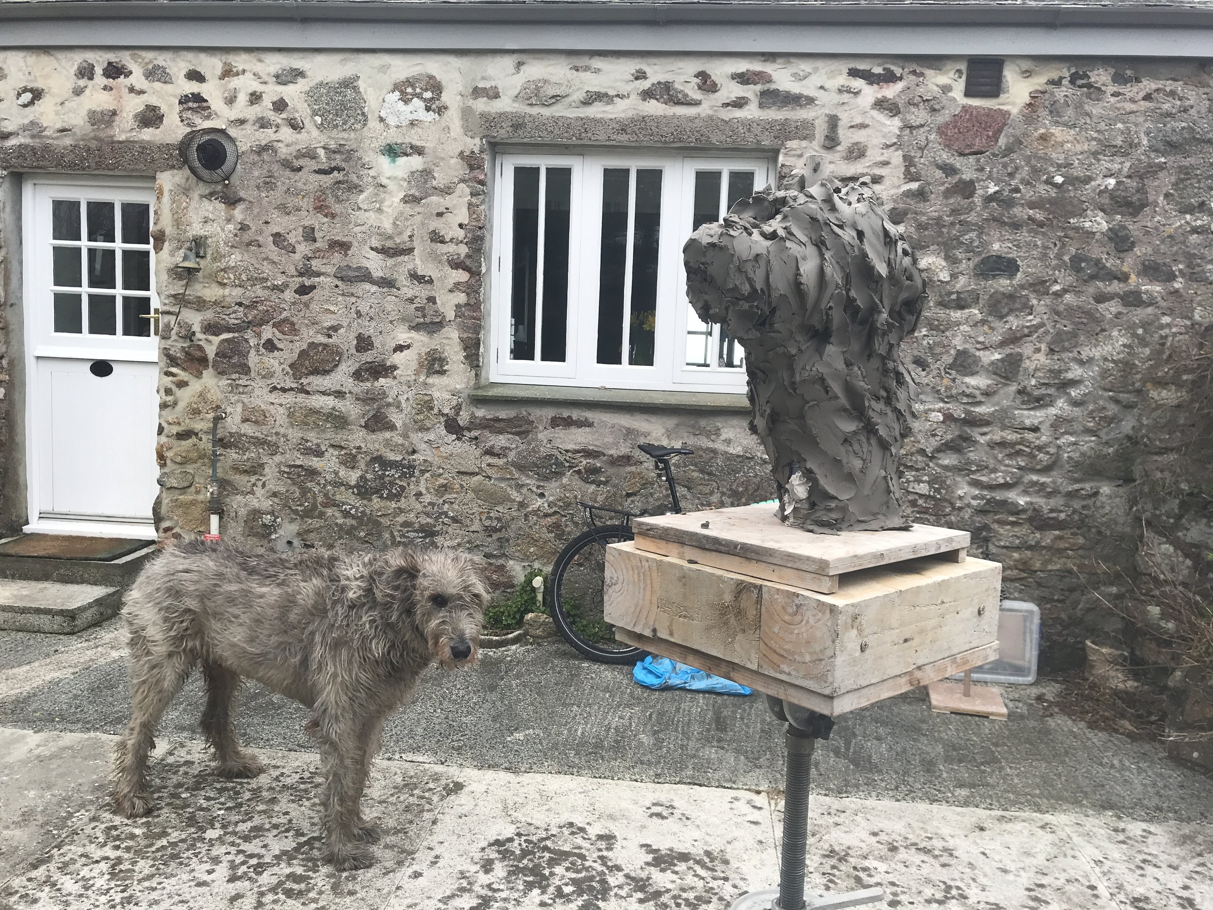 A dog standing next to a wooden sculpture on a patio outside a stone house with a white door and window.