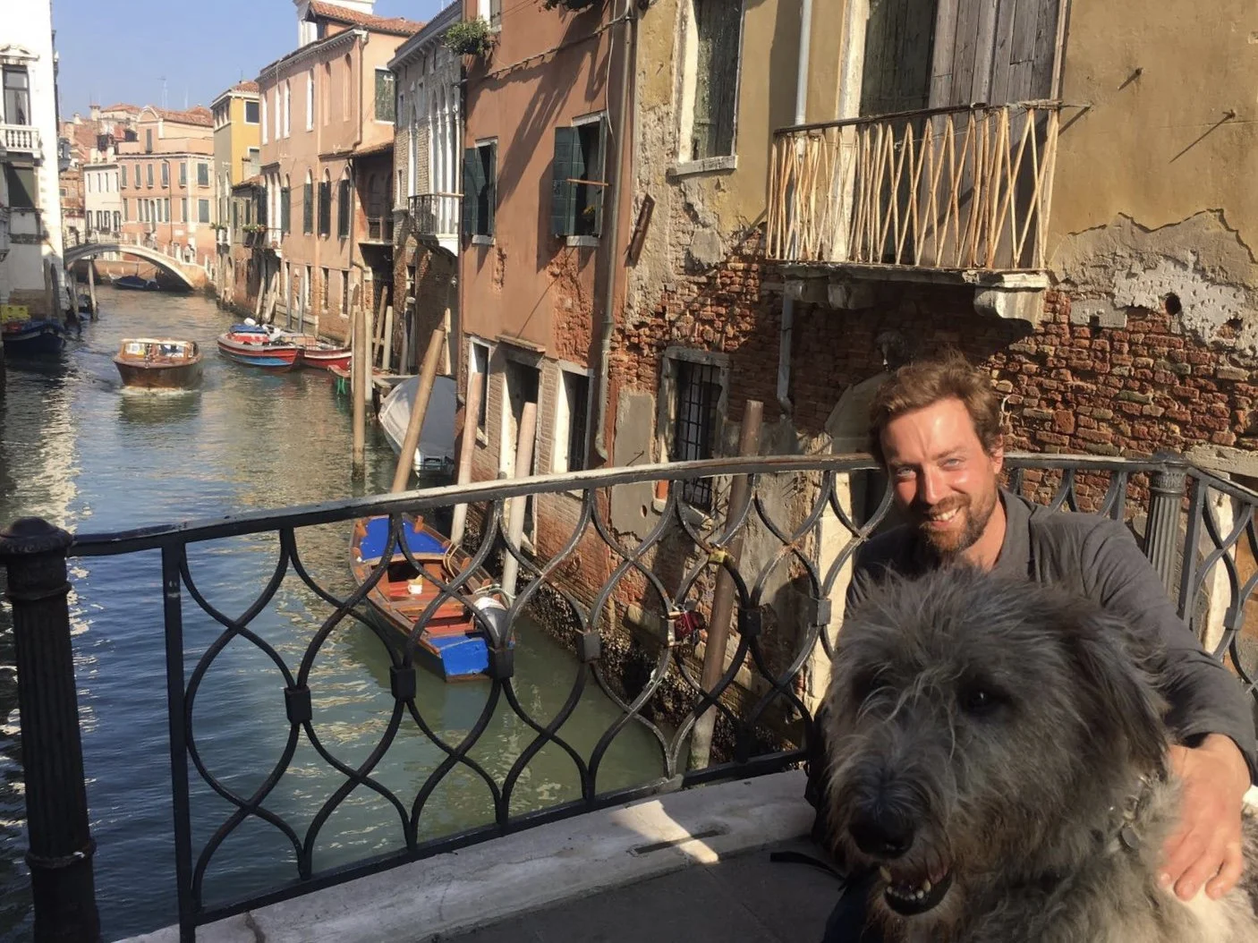 A man with a beard and a gray shirt poses on a bridge with his dog in front of a canal lined with colorful buildings and boats in Venice, Italy.