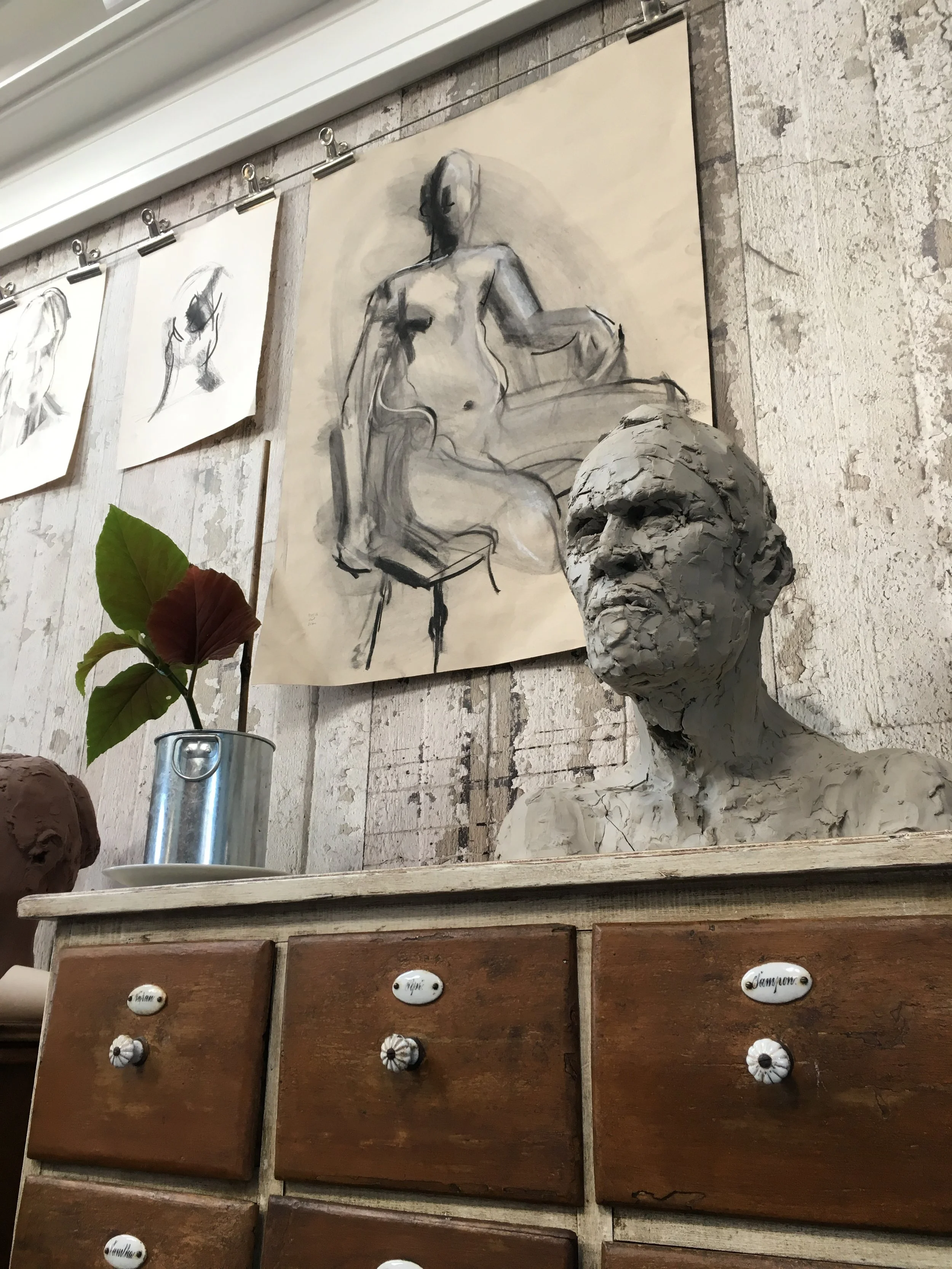 An art display with sketches of the human figure and a clay bust sculpture on a wooden chest of drawers. The artwork and sculpture are against a textured wall, with a small potted plant nearby.