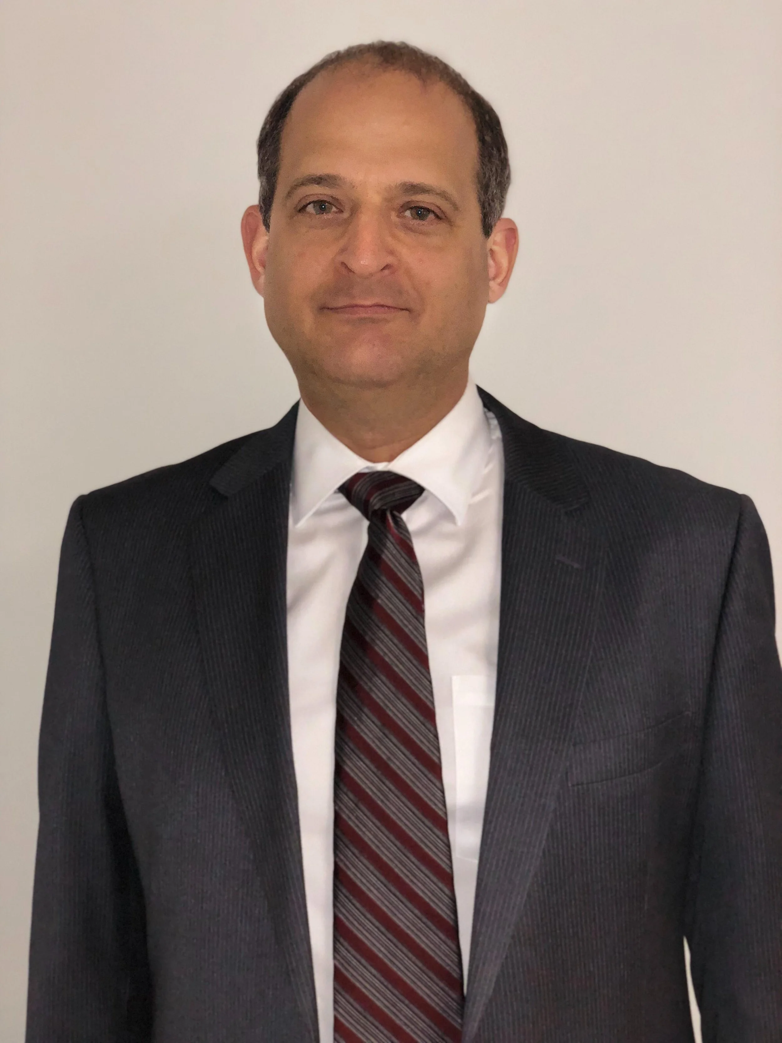 A man wearing a dark suit, white shirt, and a maroon and gray striped tie, standing against a plain white background.