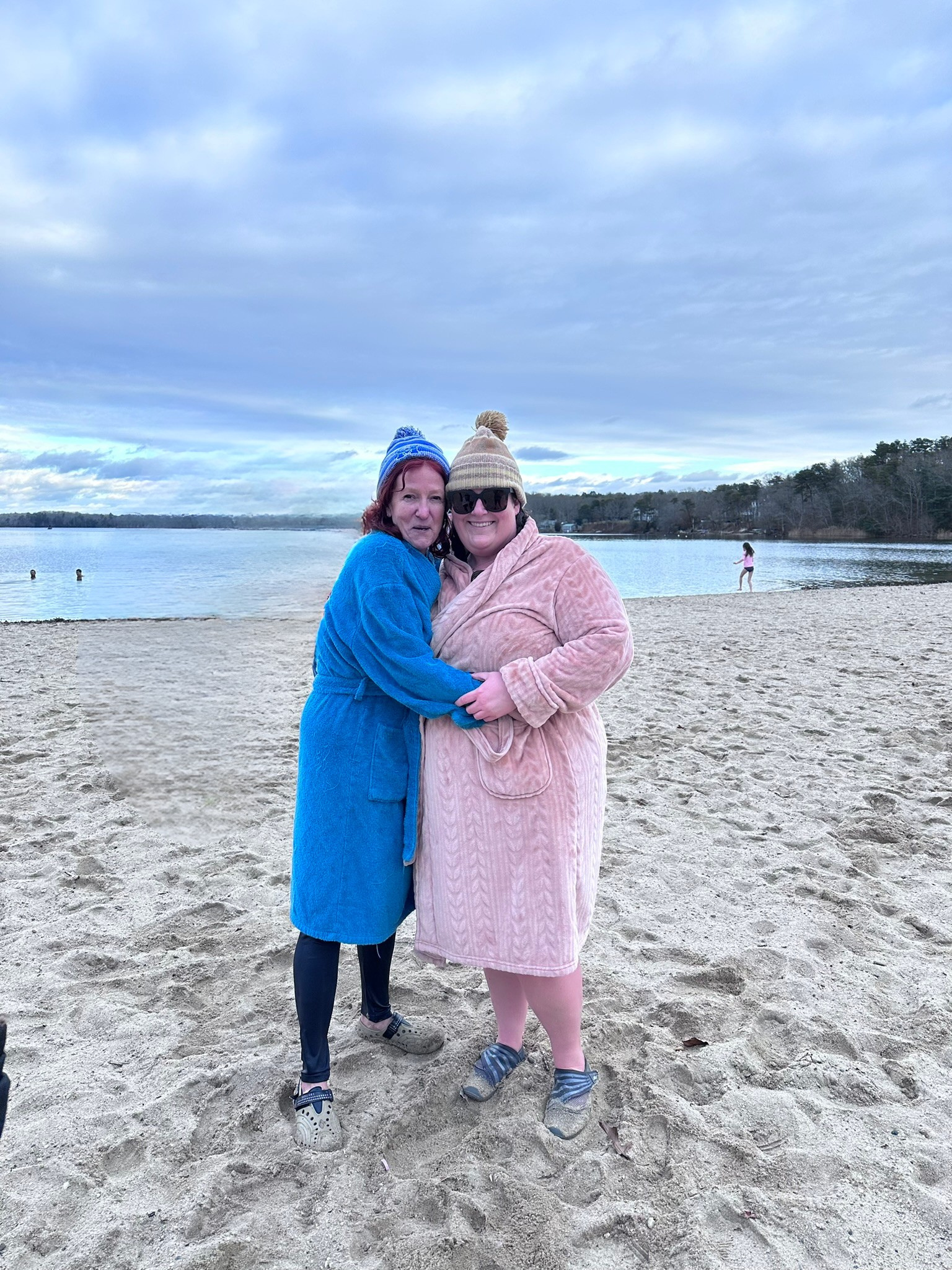 Two women standing closely together on a sandy beach near a body of water, wearing colorful winter robes and hats, with a cloudy sky overhead and a person in the background near the water.
