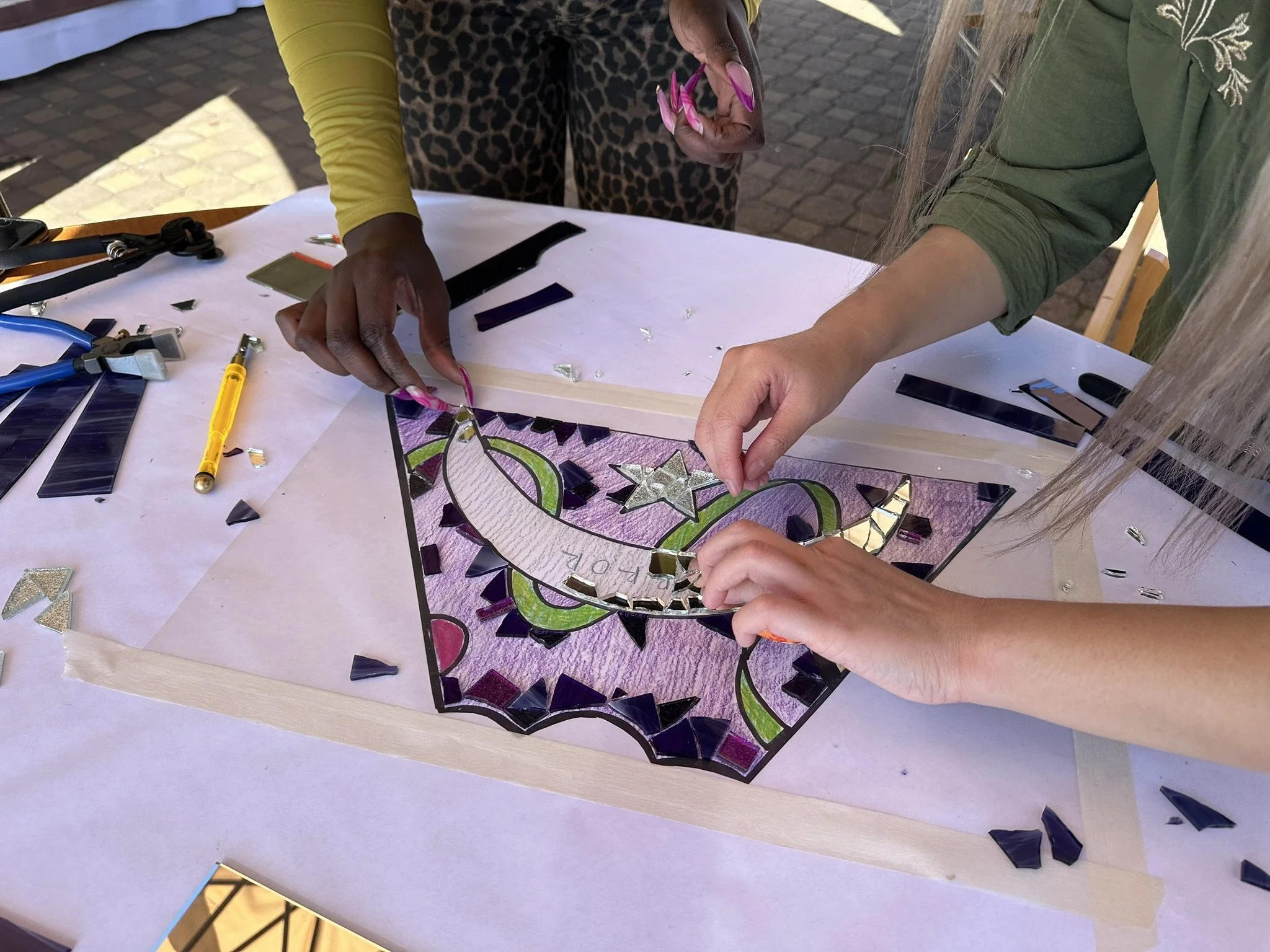 Two women working on a colorful mosaic art piece at a table lined with mosaic glass pieces, tools, and a drawing of a unicorn with stars.