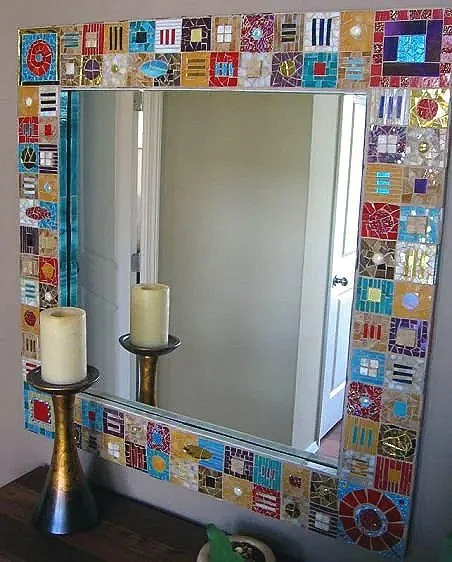 Decorative mirror with a colorful mosaic tile border, two candles on black and gold candle holders are placed in front of it, and a door reflected in the mirror.