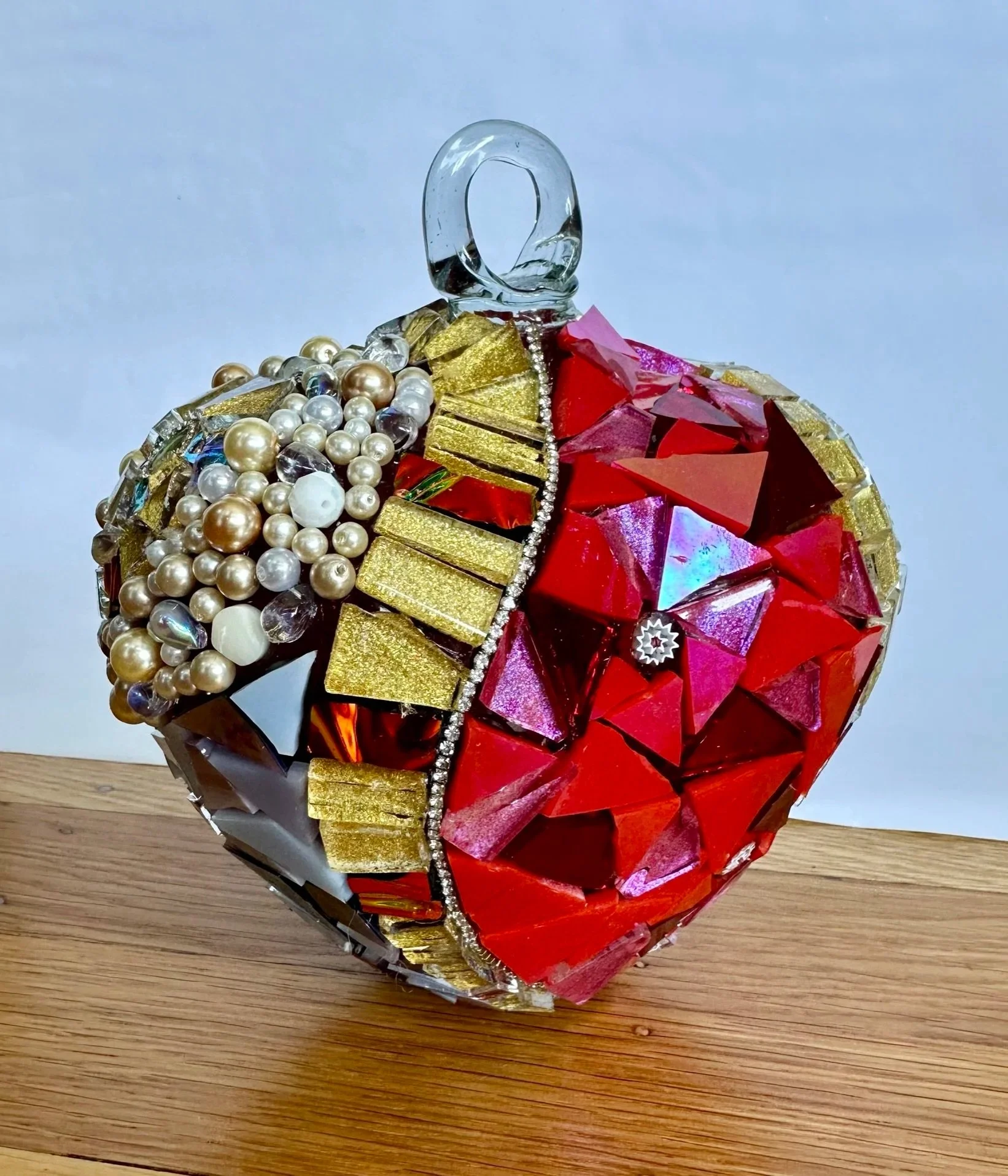 A spherical Christmas ornament decorated with beads, gold foil, and red, pink, and iridescent paper.