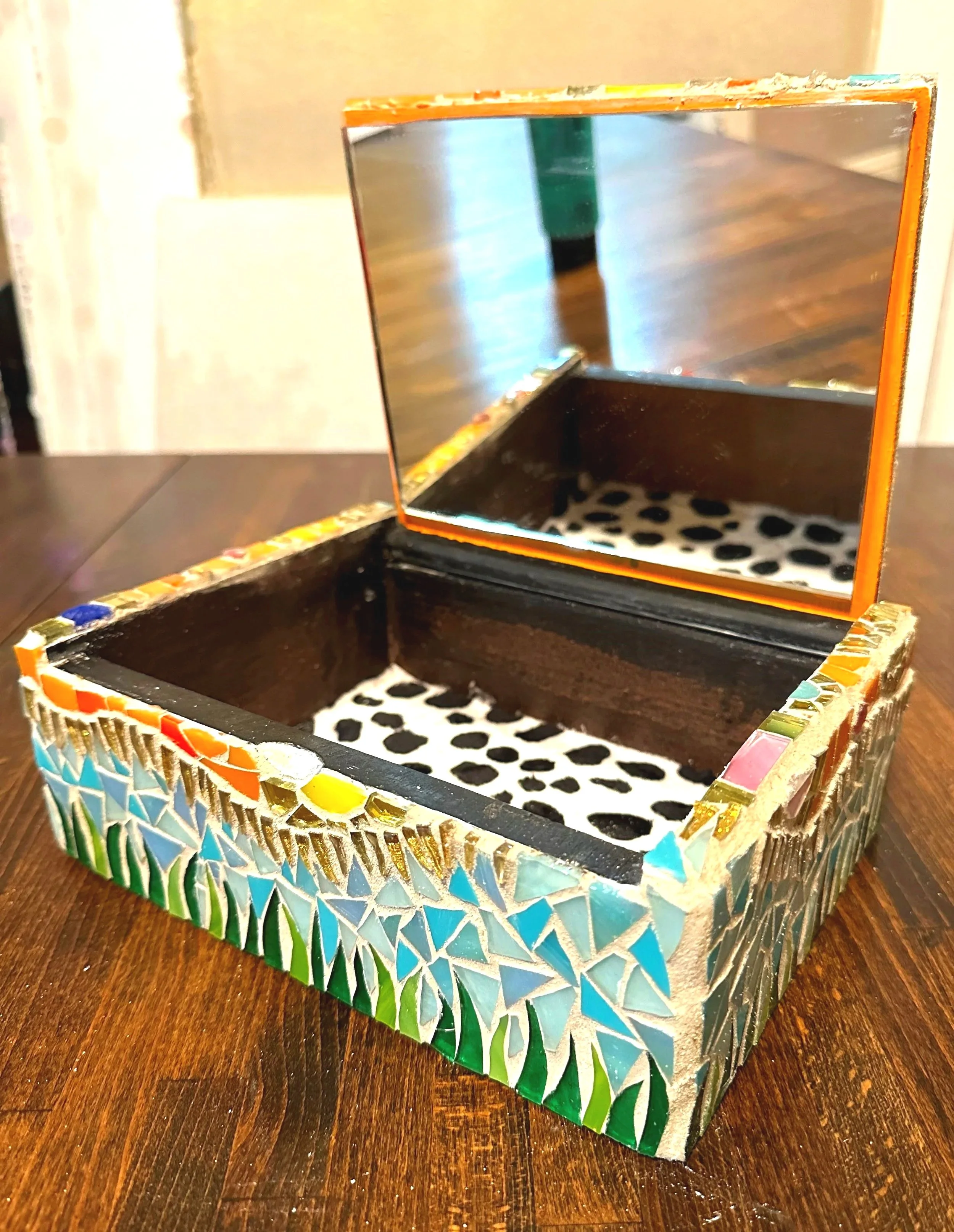A small, square mirror with a colorful, mosaic border sitting on a wooden surface. The mirror reflects part of the background, including a green bottle and a blurred wall.