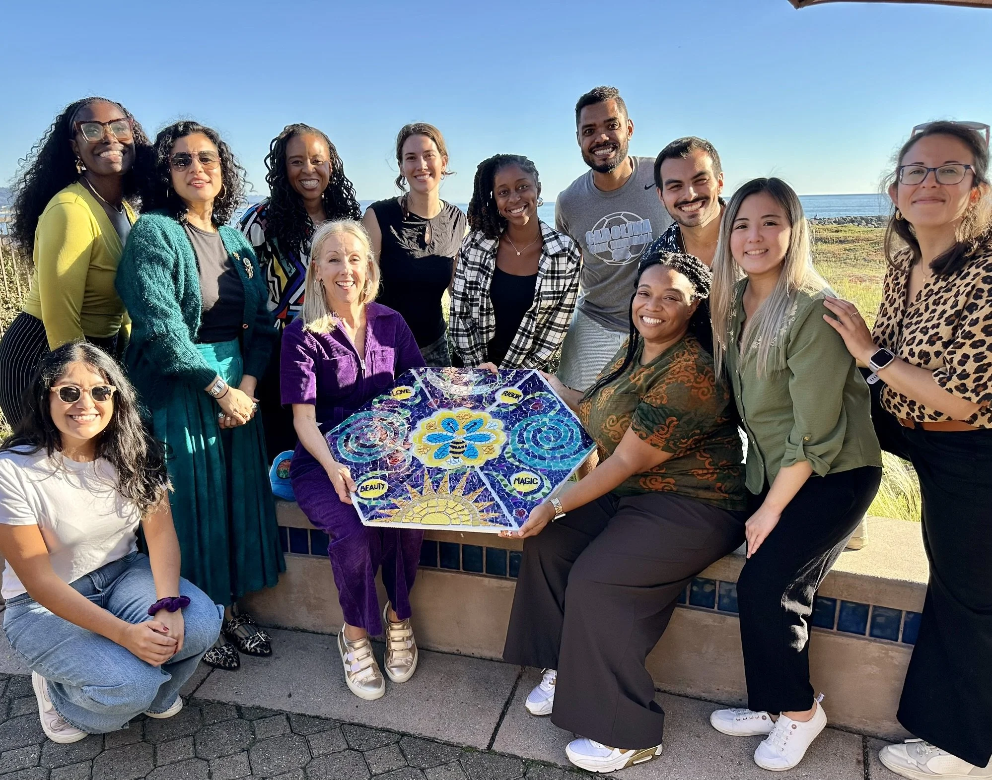 Group of diverse smiling people gathered outdoors near the ocean, some holding a colorful mosaic art piece.