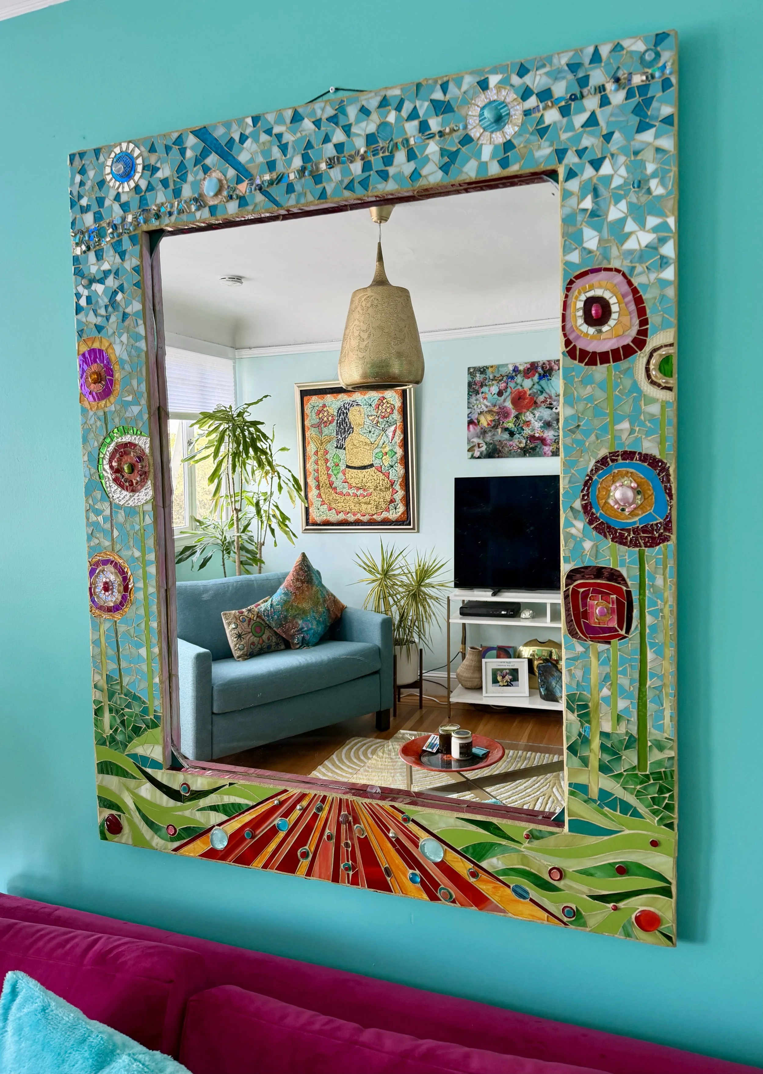 A decorated living room visible through a large, colorful mosaic mirror frame on a teal wall. The mirror reflects a light blue sofa with patterned pillows, a framed artwork of a woman, plants, a television, and a podcast playing on the table.