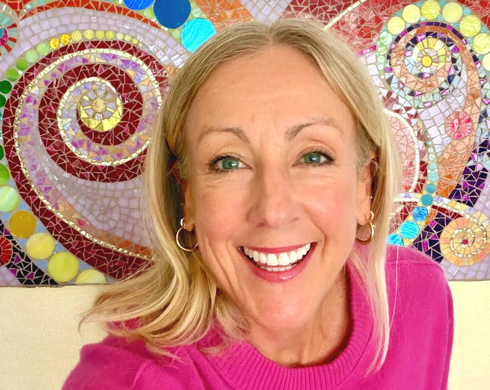 Erin Rogers with blonde hair, blue eyes, and a broad smile, wearing a pink sweater, standing in front of a colorful mosaic wall with swirls and circles made of small, shiny tiles.