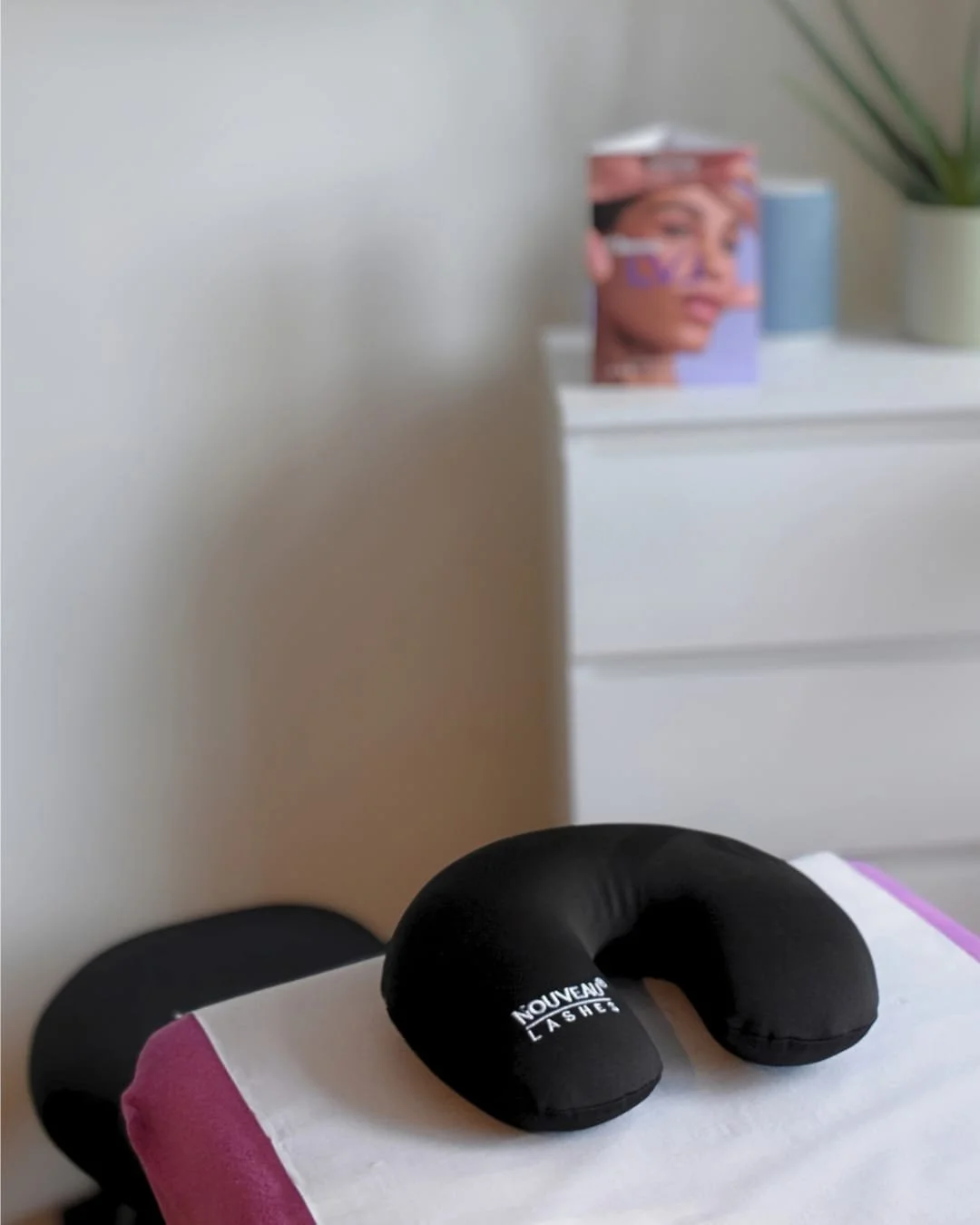 A black travel neck pillow with the words 'NOUVEAU LASHE' printed on it, resting on a pink and white surface. In the background, there is a white cabinet with a magazine or book featuring a woman's face, and a potted plant on top.