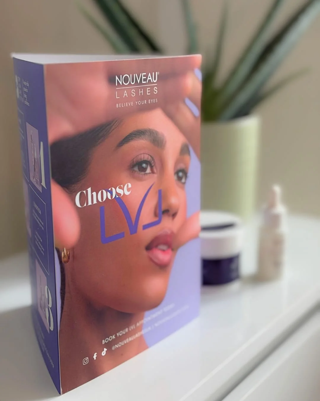 Packaging box for Nouveau Lashes with a woman's face and eye, promoting eyelash products, on a white surface with small jars and bottles in the background.
