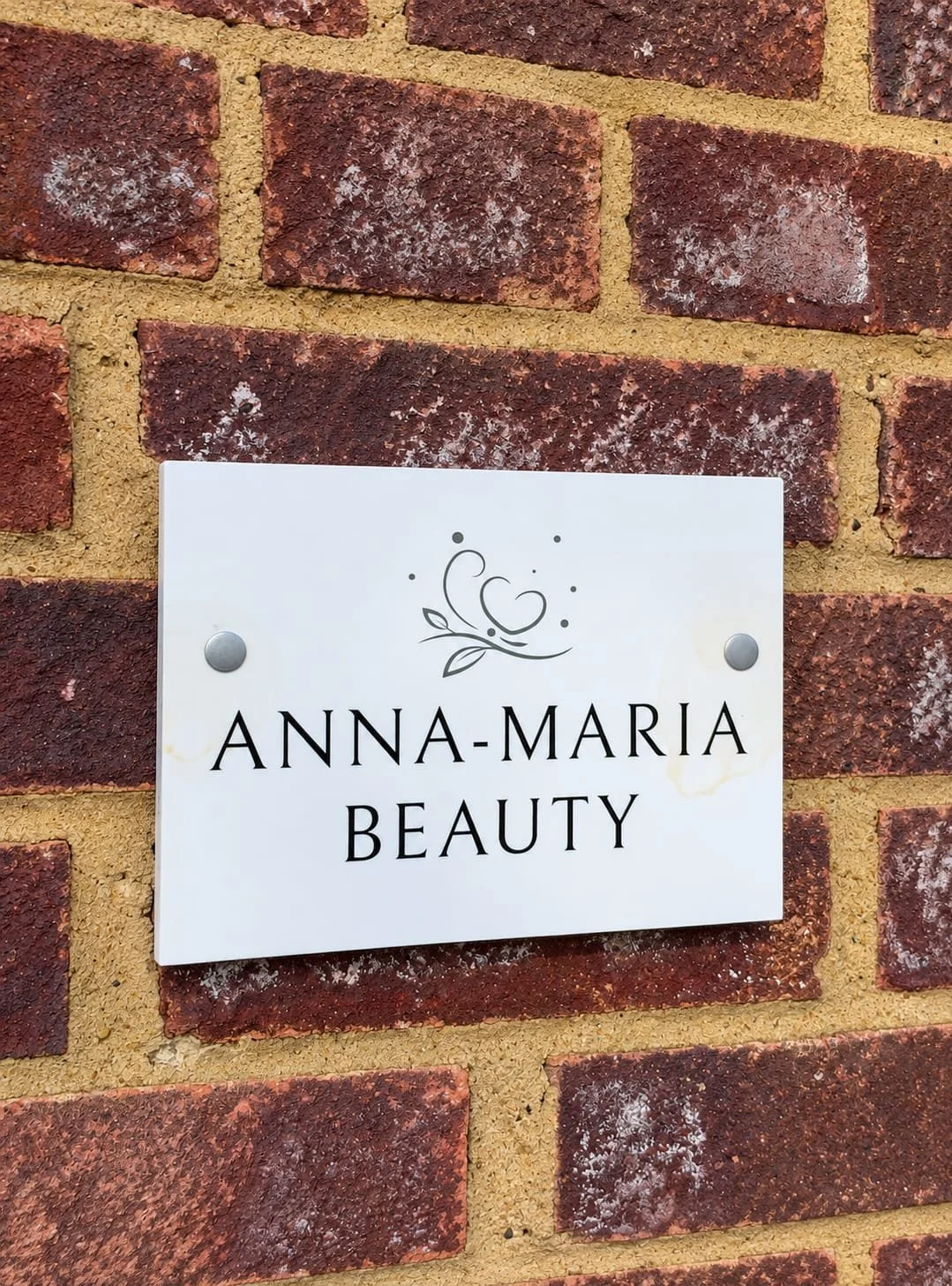 A white sign with black text reading 'Anna-Maria Beauty' mounted on a brick wall.