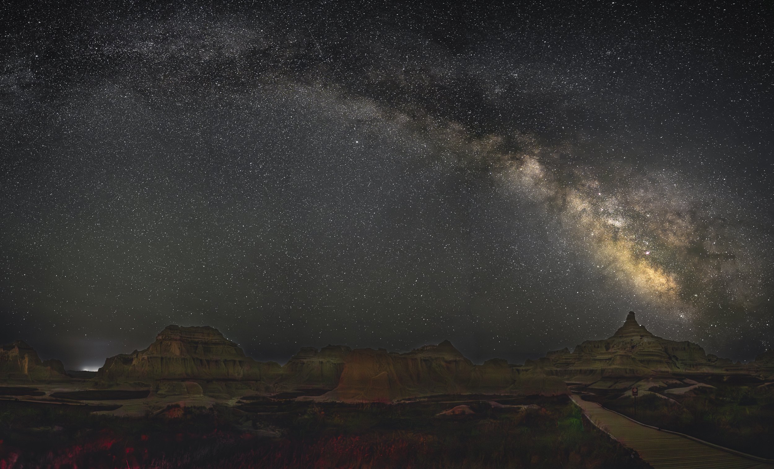 A Panoramic night sky filled with stars and the Milky Way galaxy over the South Dakota Badlands rock formations and a walking trail.