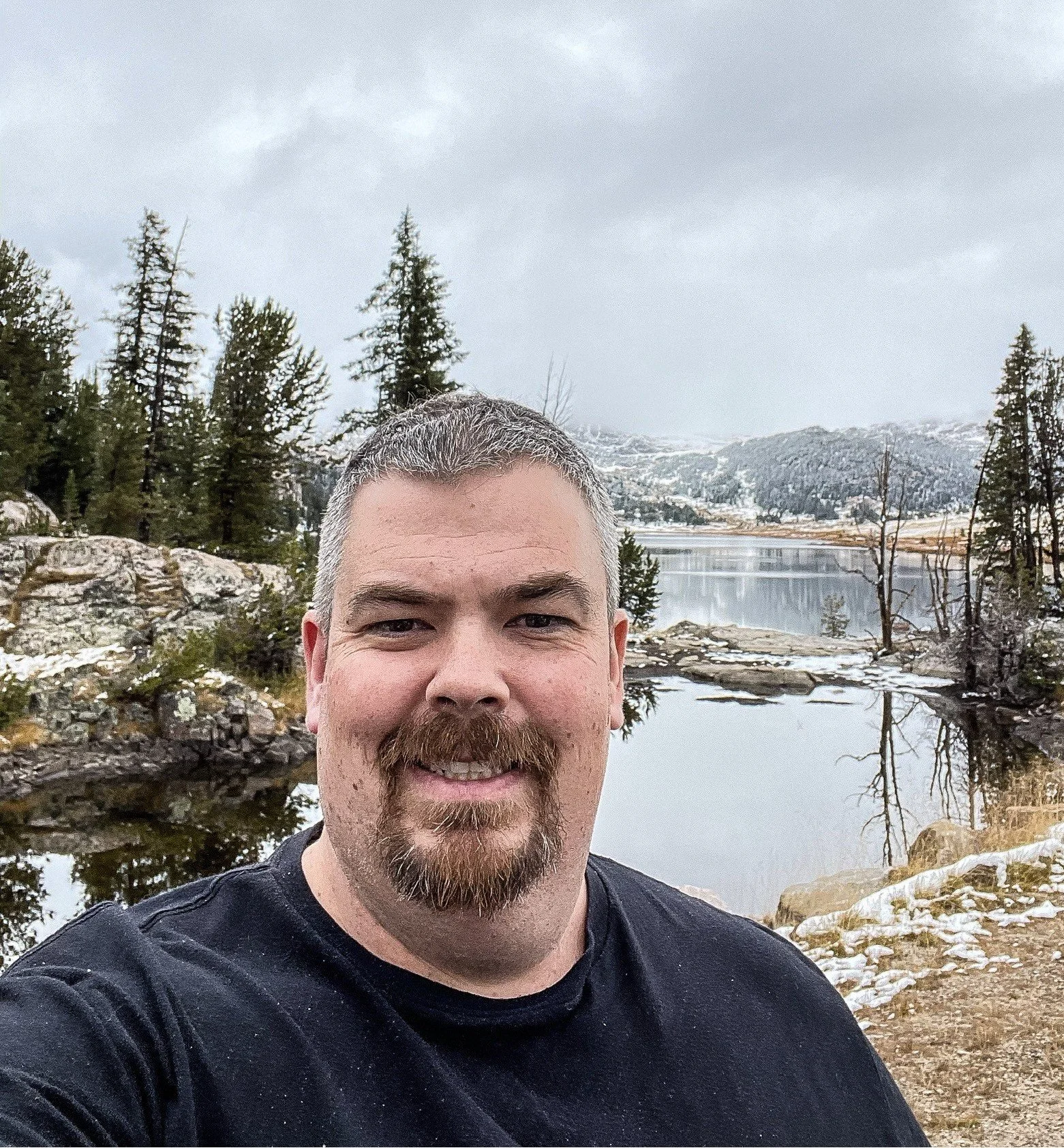 A man with short gray hair and a beard smiling outdoors near a mountain lake with trees and snow-covered mountains in the background.