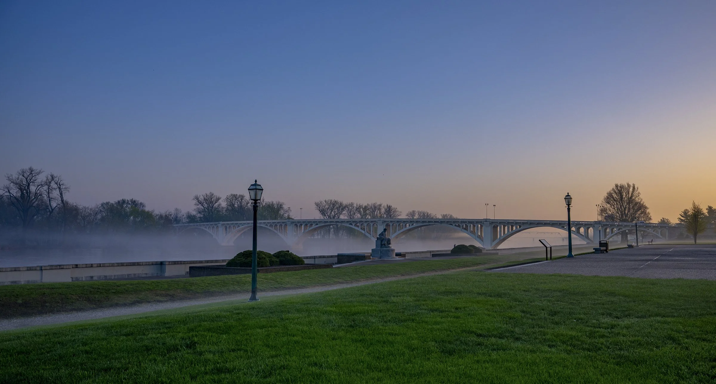 A scenic view of a park at dawn with a bridge in the background, fog over the water, and street lamps along a walkway.