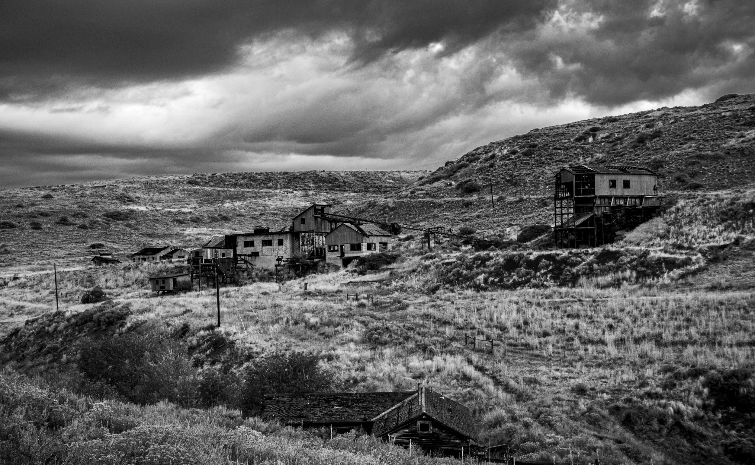 A black and white photograph of a rural landscape with old, dilapidated buildings and hills under a dark, cloudy sky.