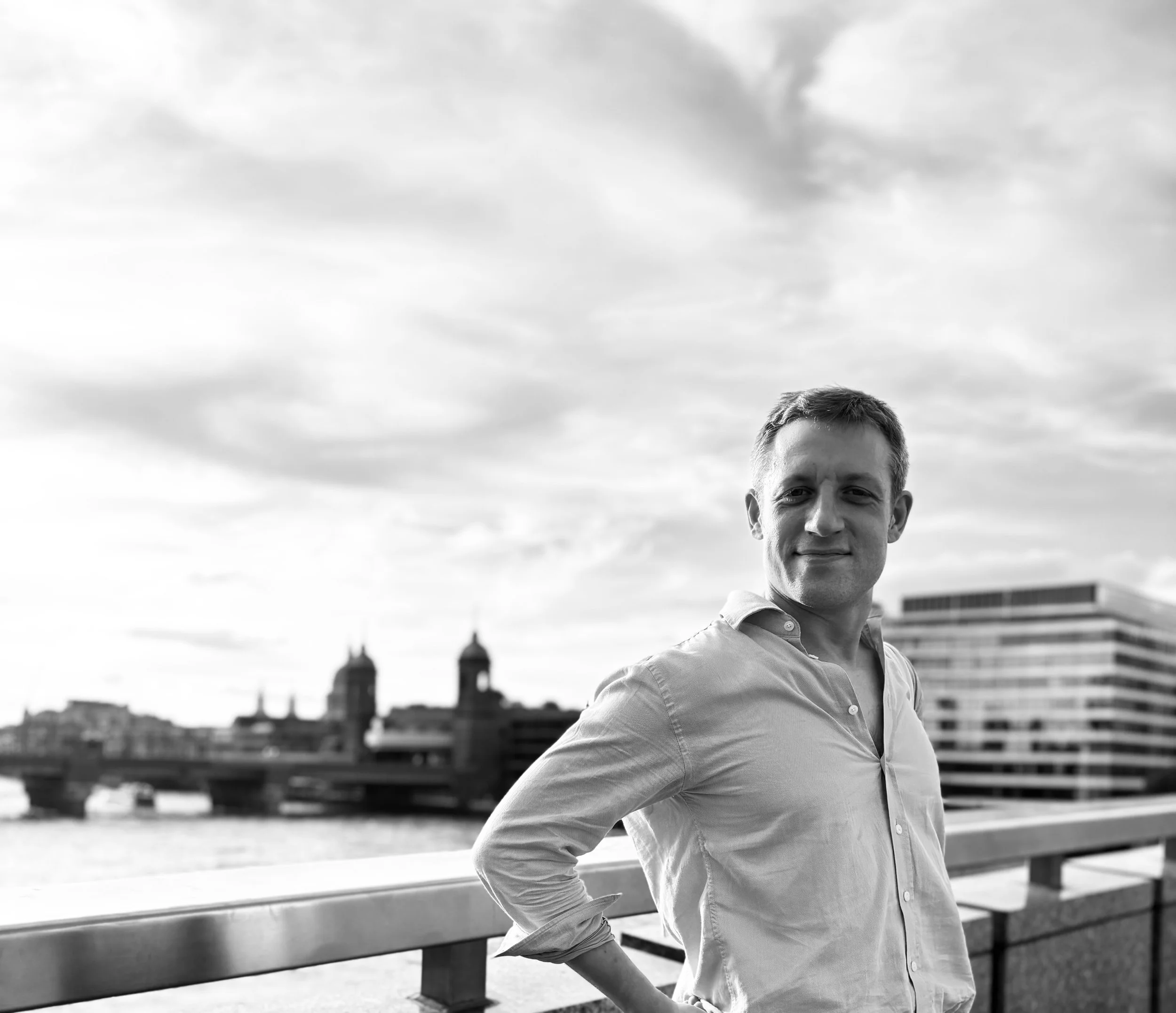 A man wearing a light-colored shirt standing outdoors on a bridge with a city skyline and cloudy sky in the background.