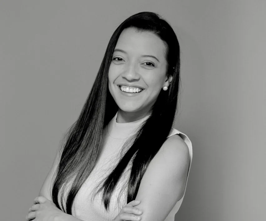 Black and white portrait of a smiling woman with long straight hair, wearing a sleeveless top and pearl earrings, standing against a plain background.