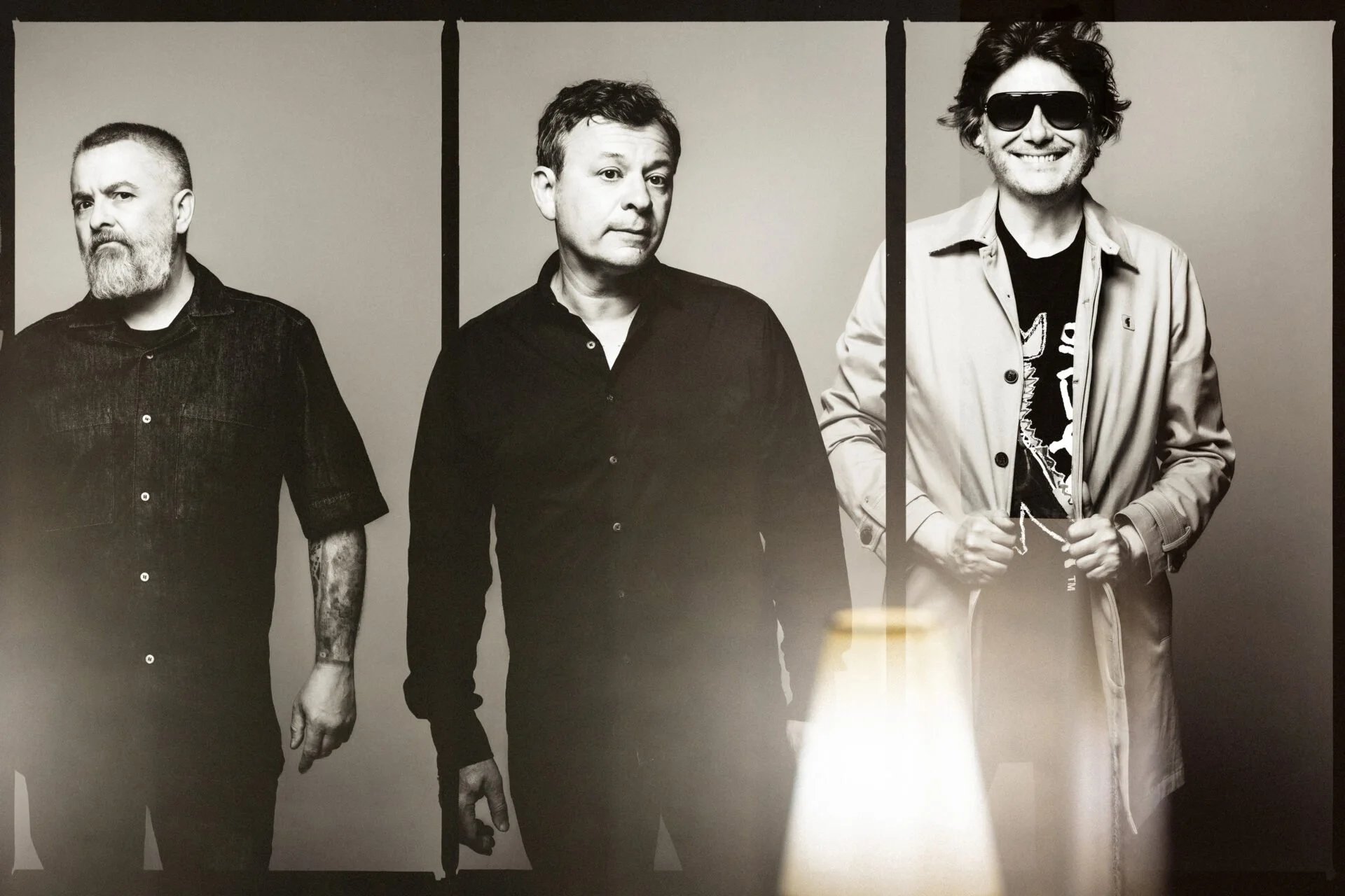 Black and white photograph of three men, Manic Street Preachers, standing side by side against a plain background. The man on the right is wearing sunglasses and smiling, while the other two have neutral expressions.