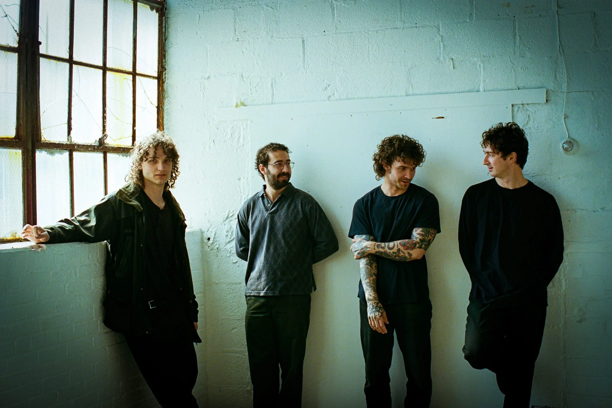 Four men with curly hair standing in a row against a white brick wall, near a large window, in an industrial-style setting.