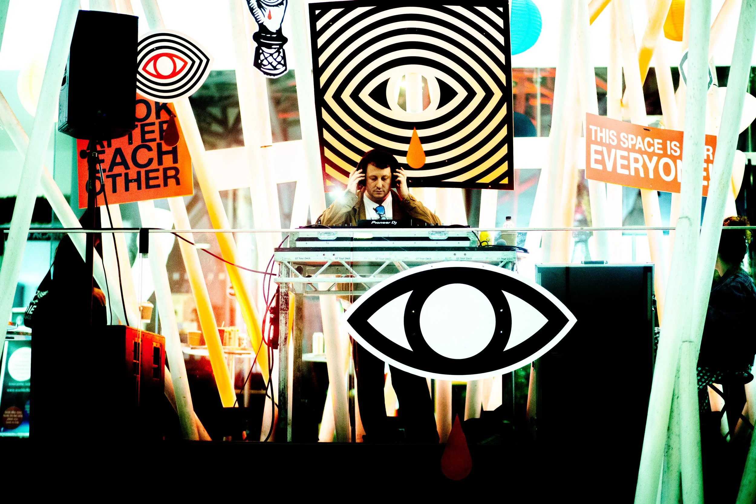 A man wearing headphones is DJing at an event with colorful, abstract and symbolic artwork and signs around him, including a large eye symbol, an eye with concentric circles, and signs with slogans like 'This space is everyone' and 'Younger.' The setting appears energetic and artistic.