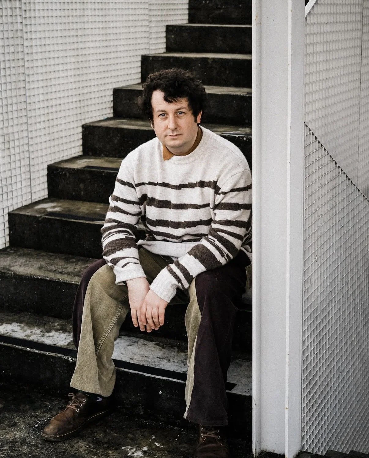 A man with dark, curly hair sitting on black stairs next to a white wall and a textured metallic wall, wearing a striped sweater and brown pants.