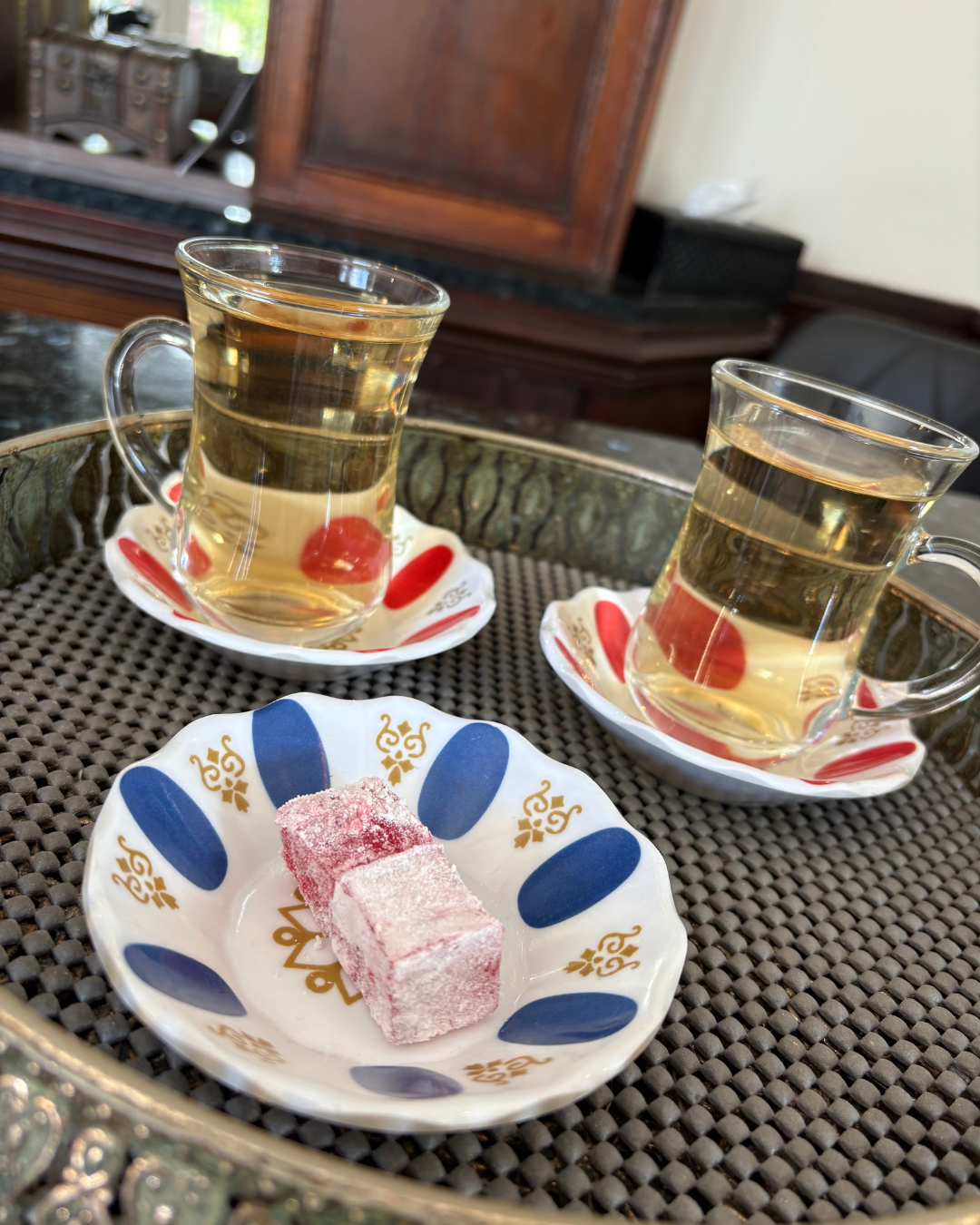 Two glasses of hot tea on decorative saucers, with a plate holding pink Turkish delight candies, set on a tray with a textured surface.