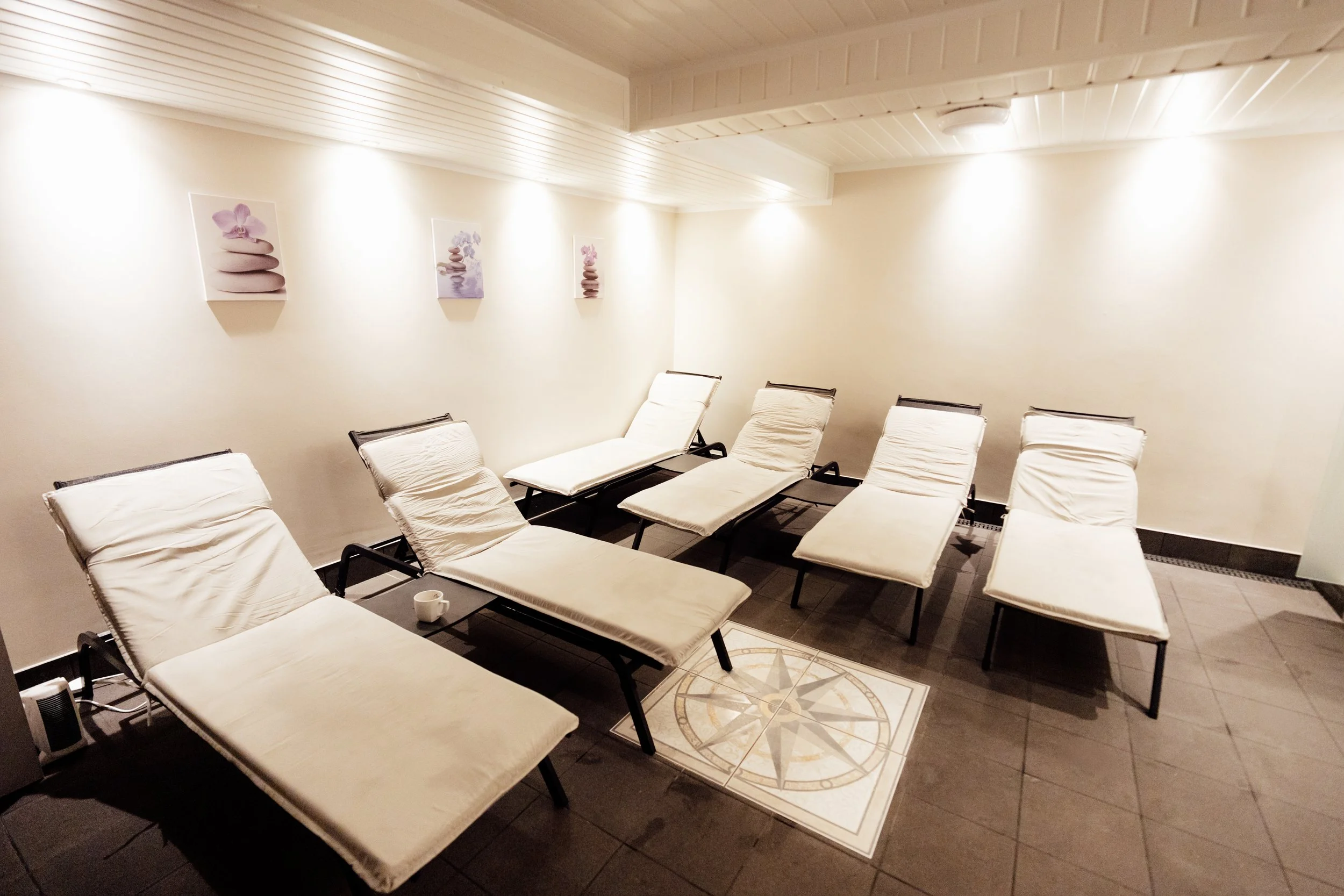 A room with white reclined chairs along the walls, three pieces of wall art featuring stacked stones and flowers, a patterned tile floor, and soft lighting.