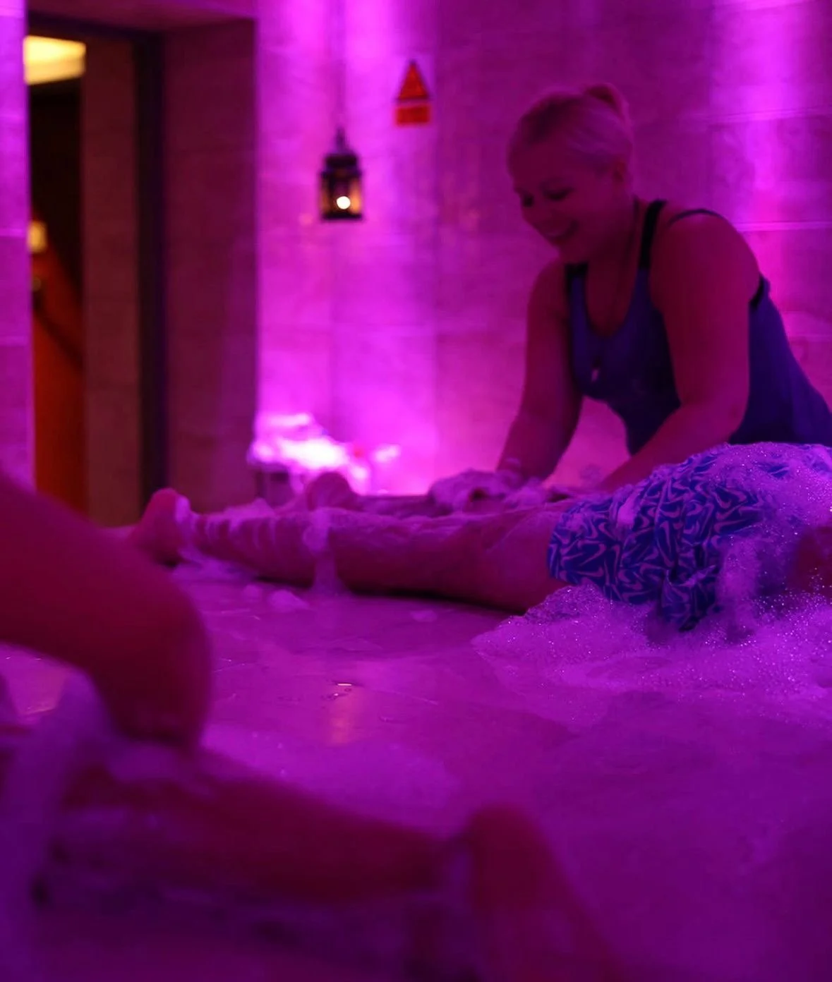 A woman giving a massage to a person lying on a table in a spa with pink and purple ambient lighting.
