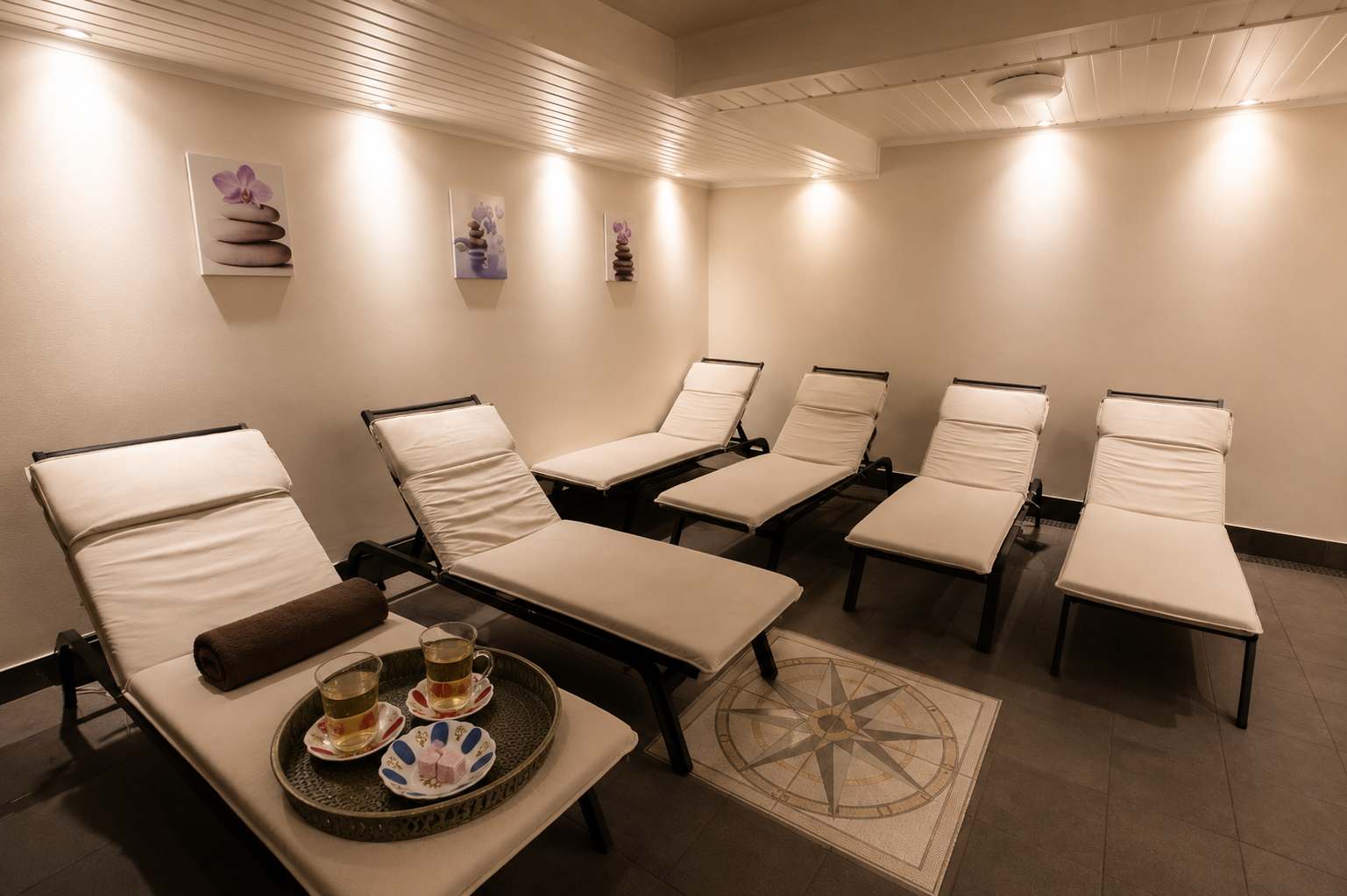 Spa relaxation room with five lounge chairs, a tray with herbal tea and sweets, and wall art of stacked stones and flowers.