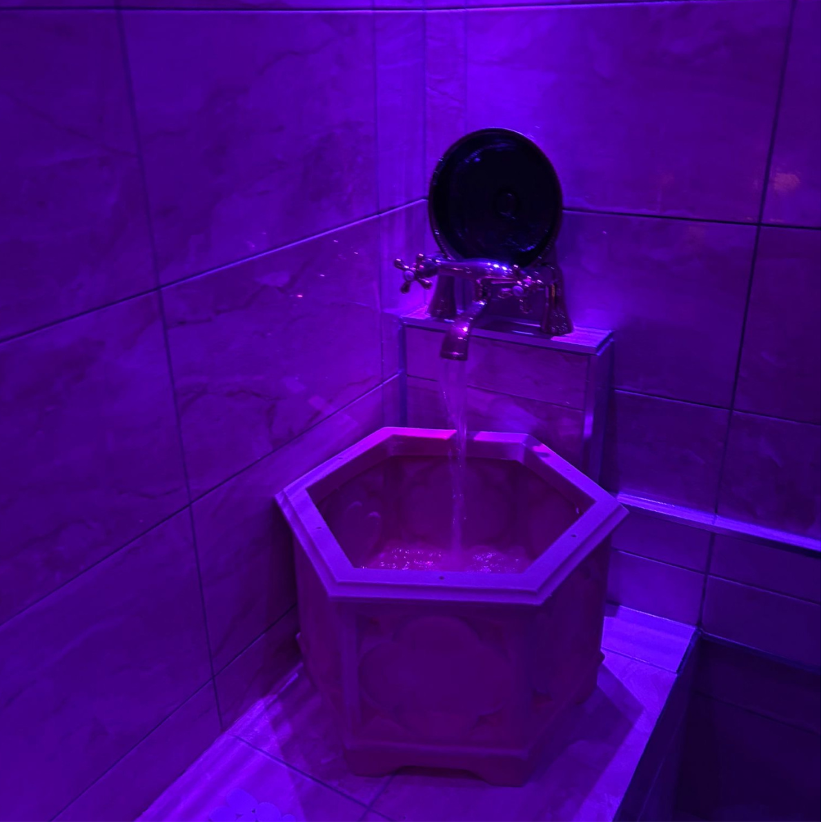A corner of a room with purple lighting, featuring a decorative fountain with water cascading from a faucet into a hexagonal basin.