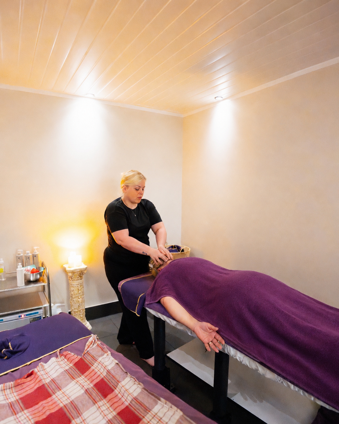 A woman giving a massage to a person lying face down on a massage table, covered by a purple towel in a wellness room.