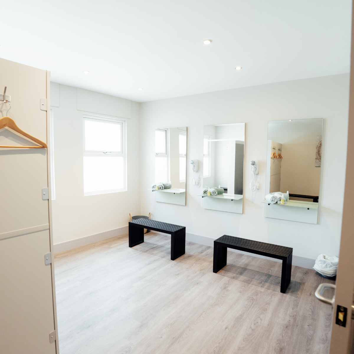 A bright, minimalist dressing room with three large vertical mirrors on the white wall, two black benches on light wood flooring, and two windows providing natural light. Towels and toiletries are placed on small white shelves beneath the mirrors.