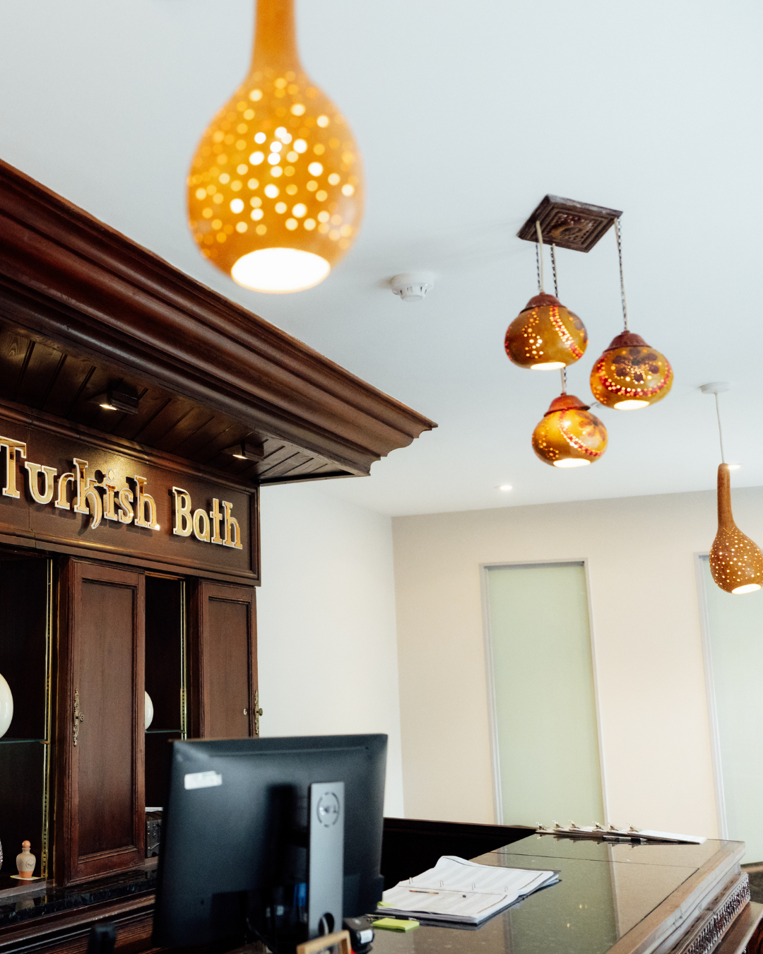 Reception area with a dark wooden desk, computer, and sign that reads 'Turkish Bath'. Hanging decorative lighting fixtures are visible on the white ceiling.