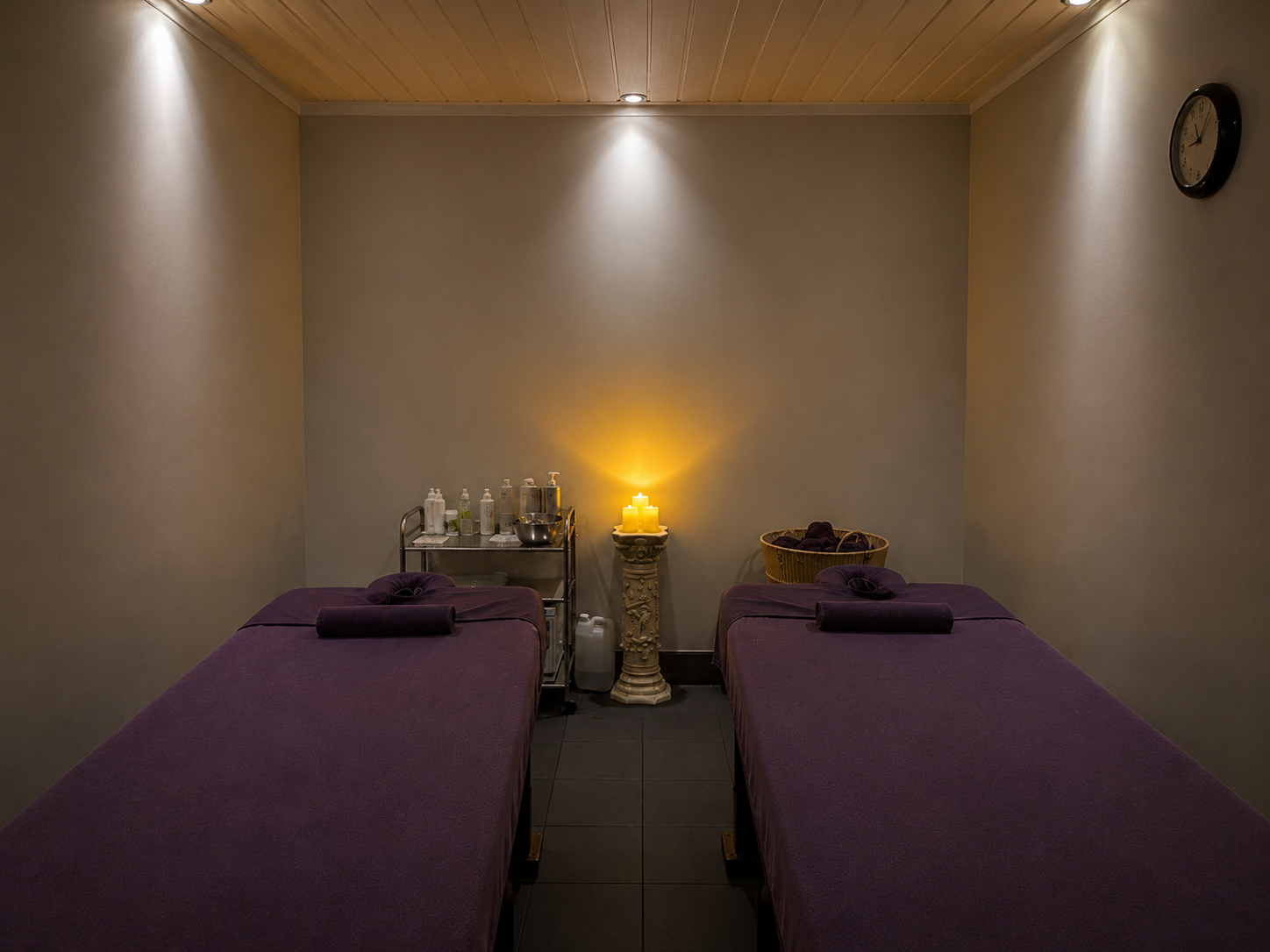 Spa treatment room with two massage tables covered in purple linens, candles, sanitizer bottles, and a clock on the wall.