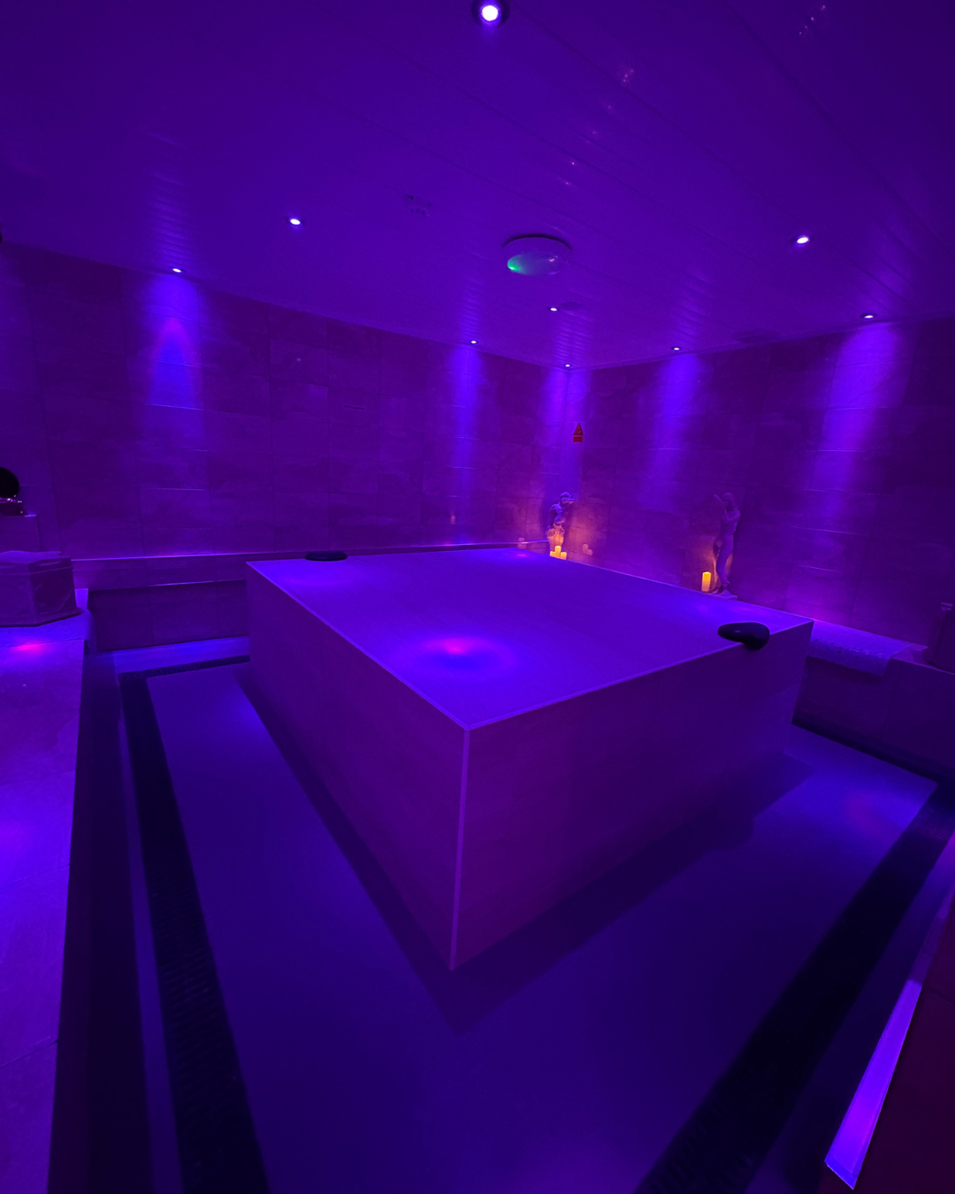 A dimly lit relaxation or spa room with purple lighting, featuring a large square hot tub in the center, statues on the walls, and candles providing soft illumination.