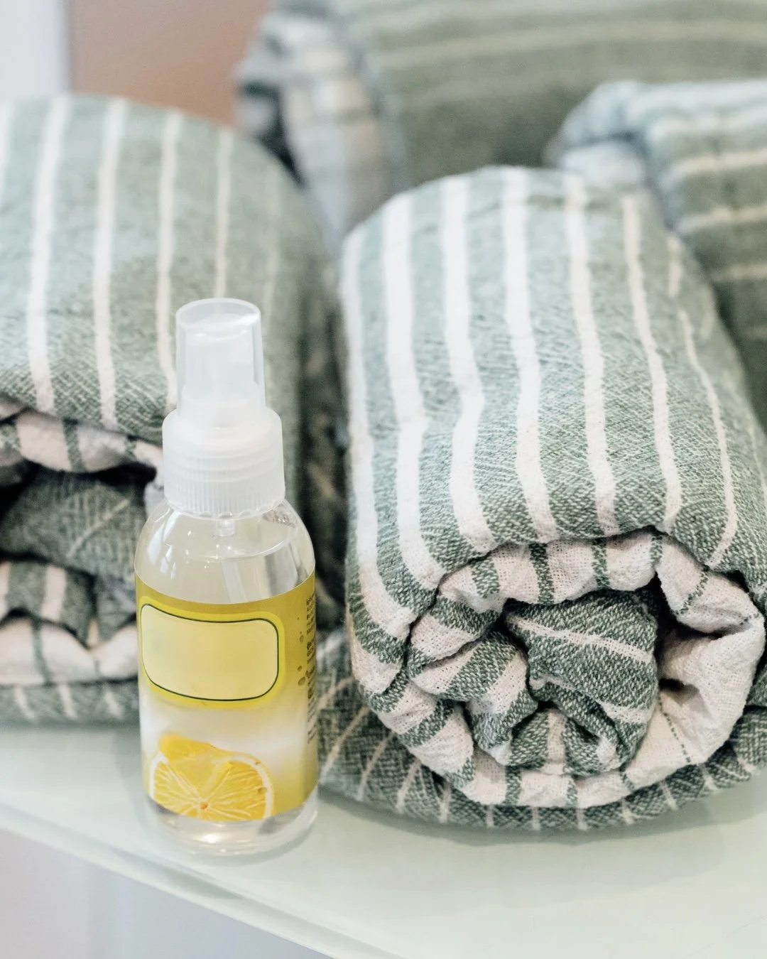 A clear spray bottle with a lemon design label sits in front of three rolled-up, green and white striped towels.