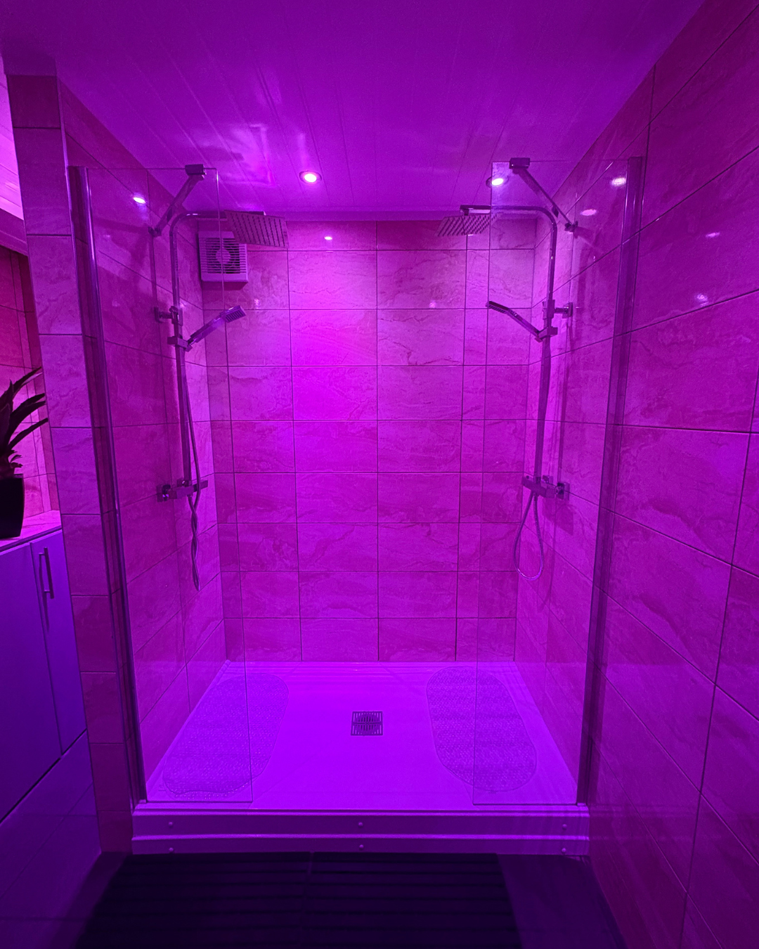 A walk-in shower with pink and purple lighting, two showerheads, and beige tiled walls.