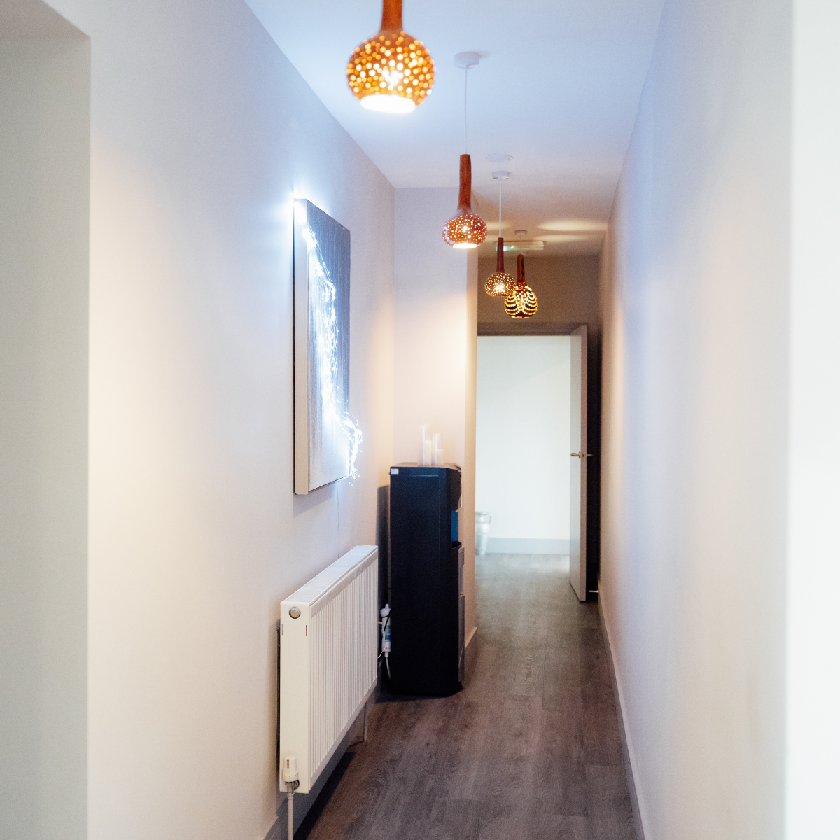 A hallway with five hanging pendant lights, a painting on the wall, a radiator, and a black speaker cabinet, leading to a bathroom with a toilet.
