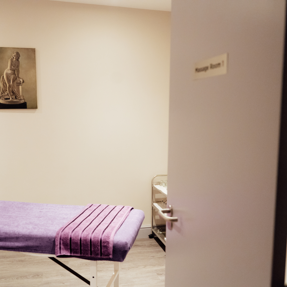 A massage room with a massage table covered with a purple towel, a small cart, and a wall with a framed picture of a seated woman statue.